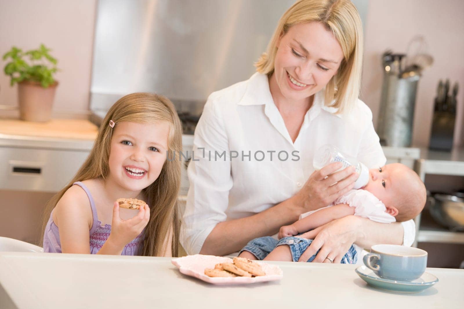 Mother feeding baby in kitchen with daughter eating cookies and smiling