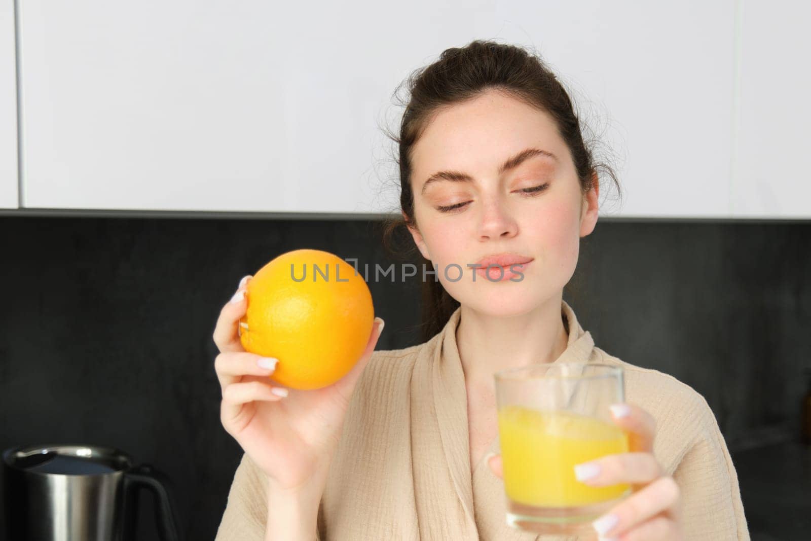 Portrait of beautiful, healthy young woman with orange in hand, drinks freshly squeezed juice from glass, posing in kitchen, enjoys her morning at home.