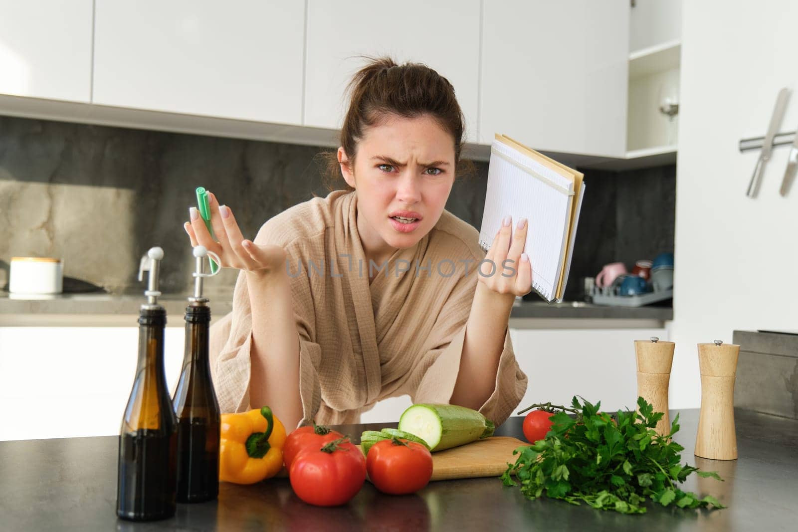 Portrait of woman cant cook, looking confused while making meal, holding recipe book, checking grocery list and staring frustrated at camera, standing near vegetables in the kitchen by Benzoix