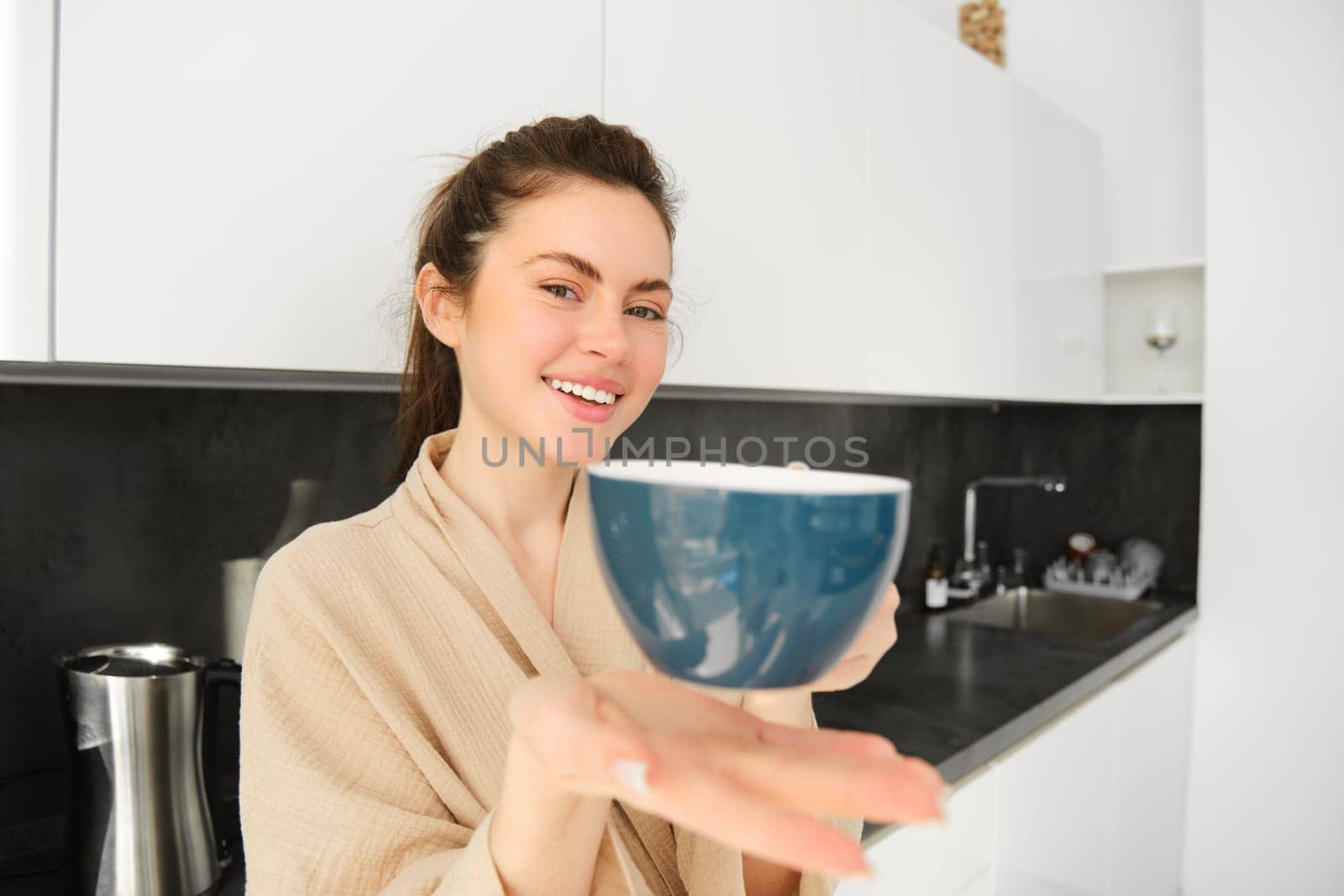 Portrait of attractive smiling woman giving you cup, offering morning coffee and looking happy, standing in the kitchen in bathrobe.