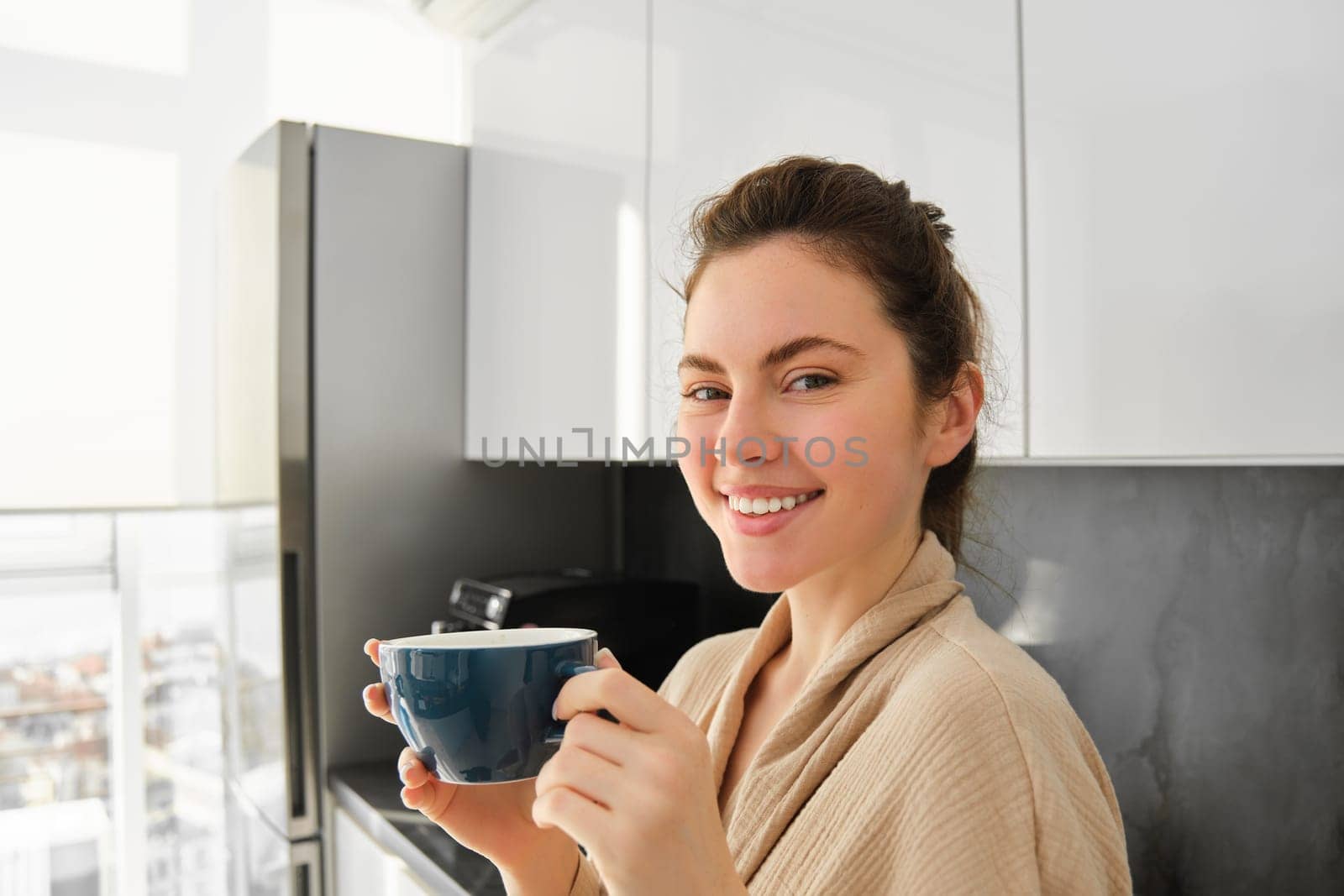 Daily routine and lifestyle. Young beautiful woman in bathrobe, standing in kitchen with cup of coffee, drinking tea, smiling and looking happy.