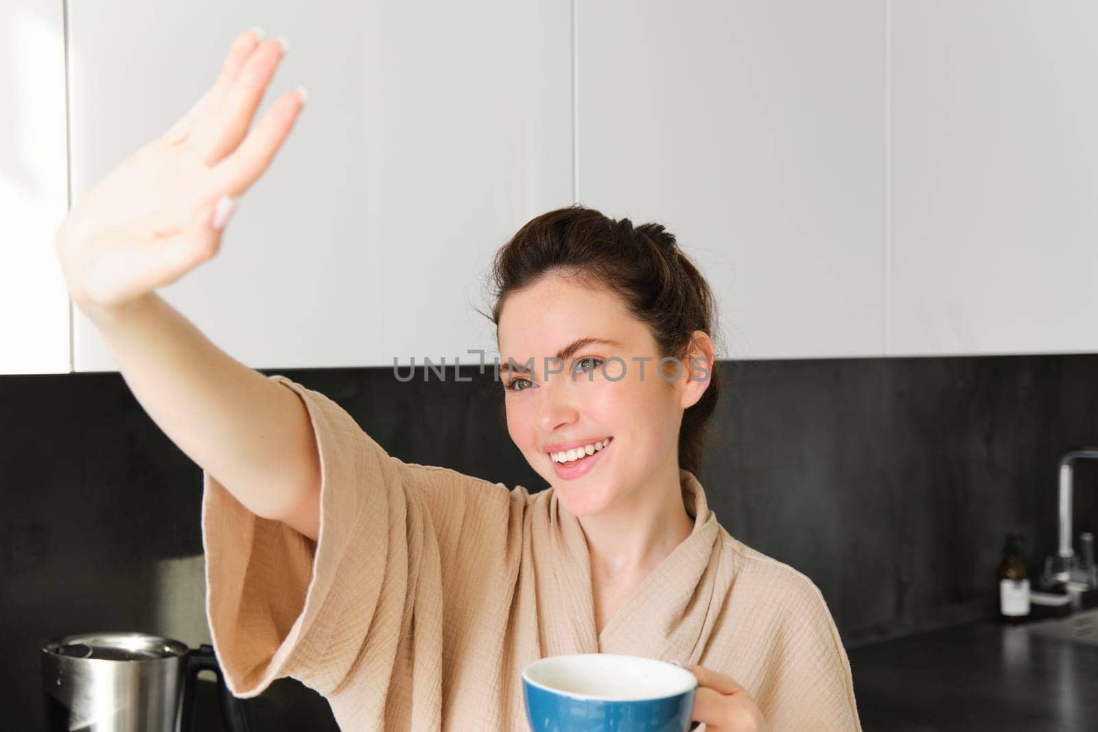 Close up portrait of carefree woman, covers her eyes from sunlight, looking out of window in the kitchen, drinking morning coffee and smiling.