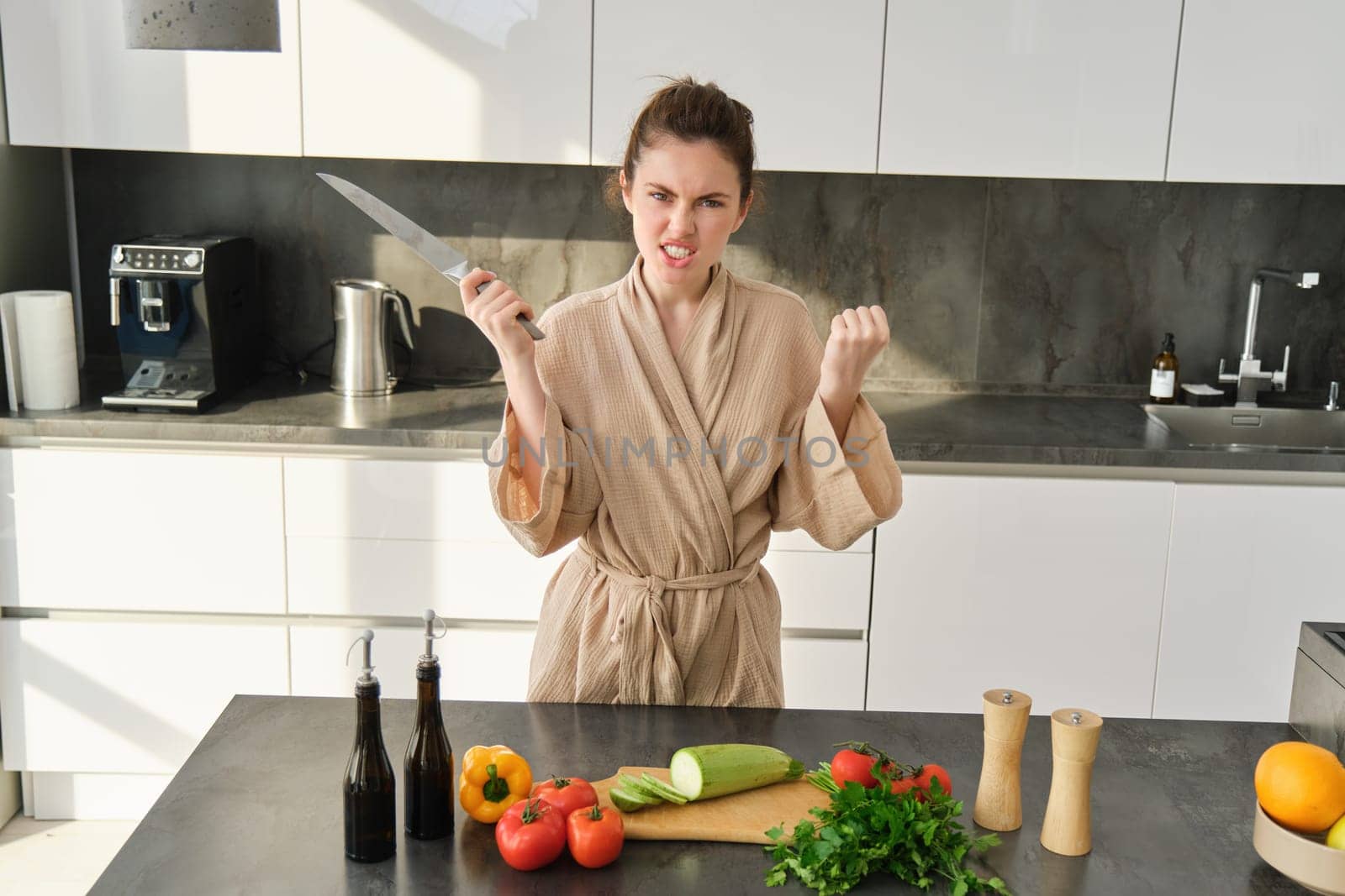 Angry brunette woman with knife, standing in the kitchen, annoyed and frustrated to cook, making upset grimace, standing in bathrobe by Benzoix