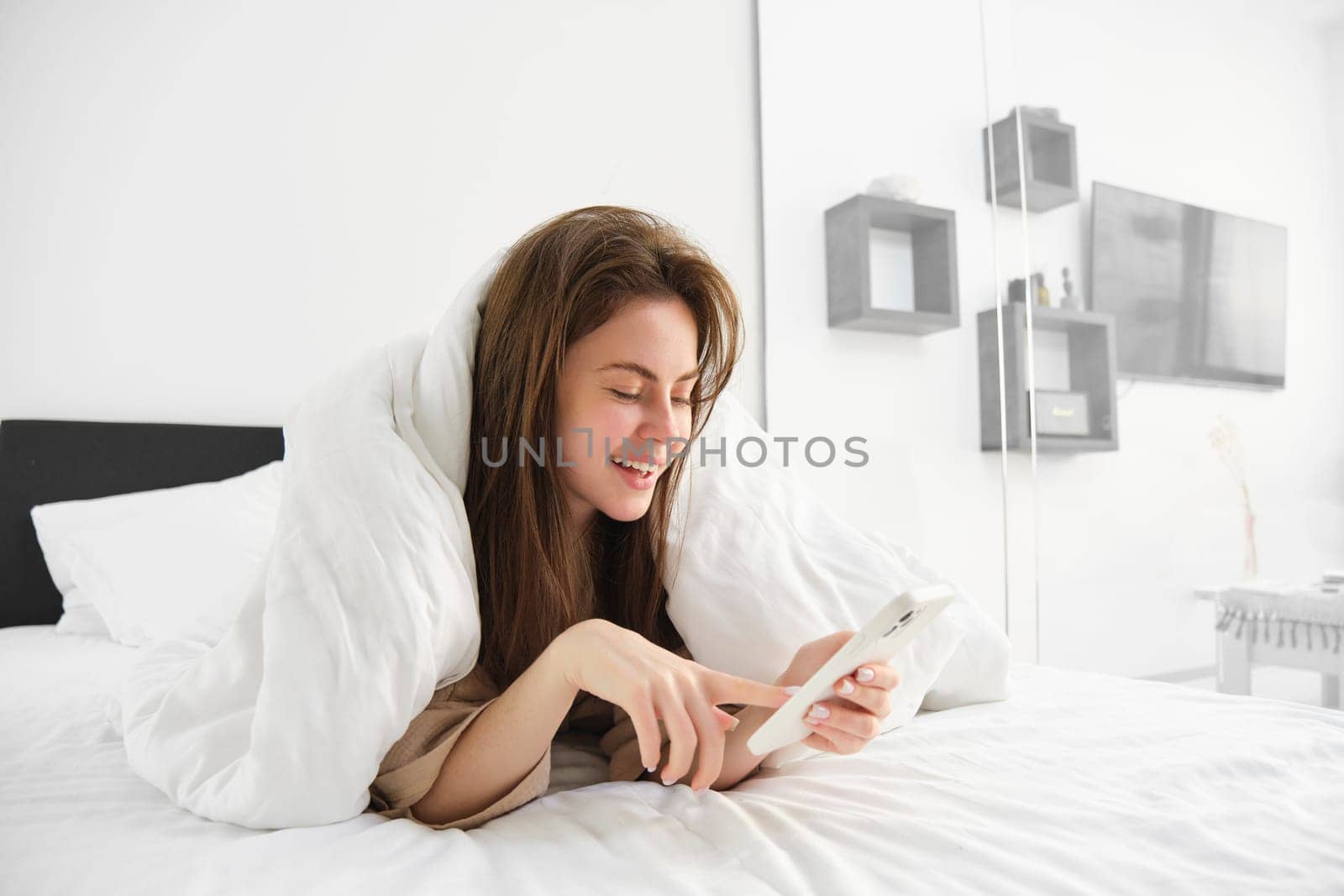 Portrait of beautiful brunette woman with smartphone, relaxing in her bed under blankets and smiling, checking social media feed on mobile phone app, wrap herself in duvet and enjoying lazy morning.