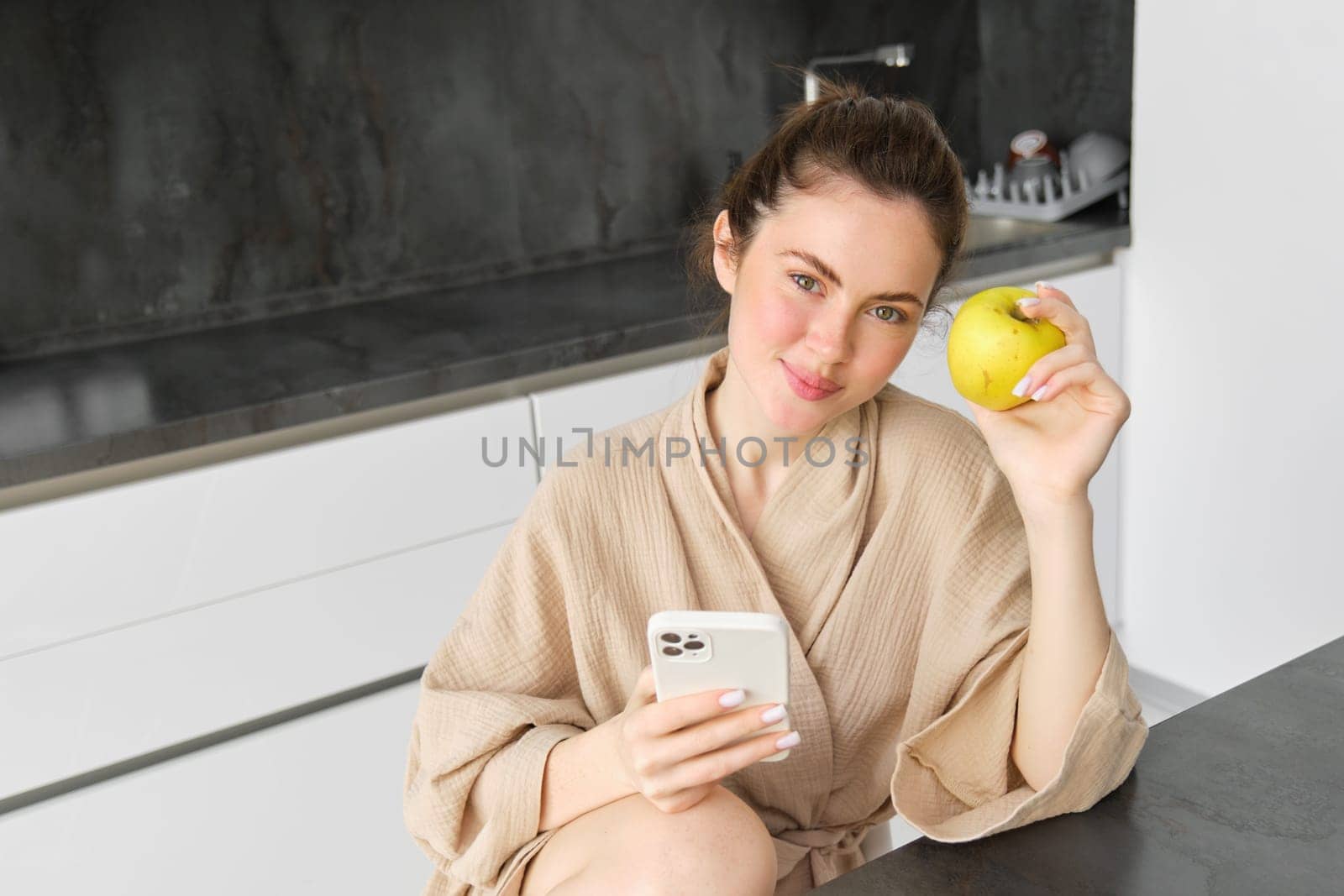 Image of attractive young woman in bathrobe, sits in kitchen, eats green apple and looks at mobile phone, uses smartphone app, orders food on application.