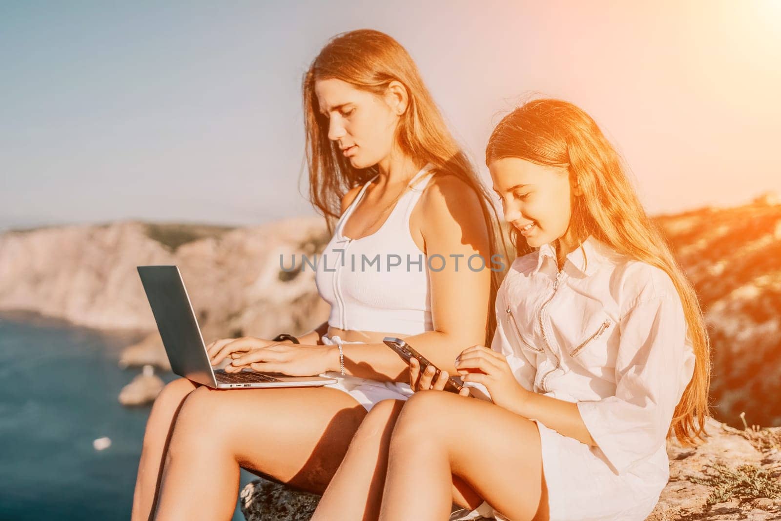 Digital nomad, Business woman working on laptop by the sea with daughter. Pretty lady typing on computer by the sea at sunset, makes a business transaction online. Freelance, remote work on vacation by panophotograph