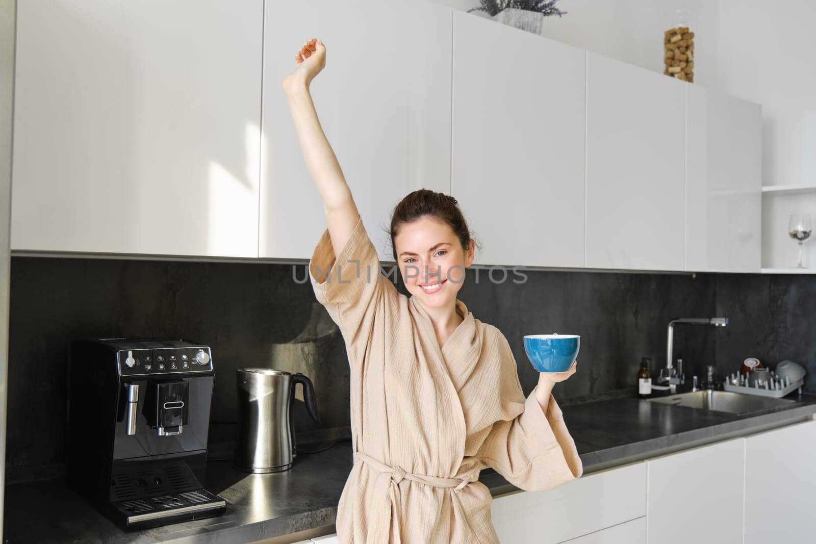 Portrait of carefree brunette woman, dancing and stretching hands in the kitchen, drinking coffee, smiling pleased in the morning.
