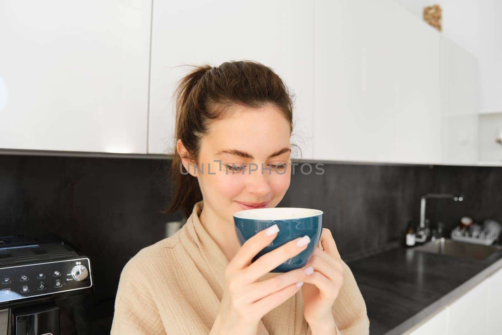 Portrait of beautiful young woman in bathrobe, drinking morning coffee and enjoying the taste, smiling pleased, standing in the kitchen.