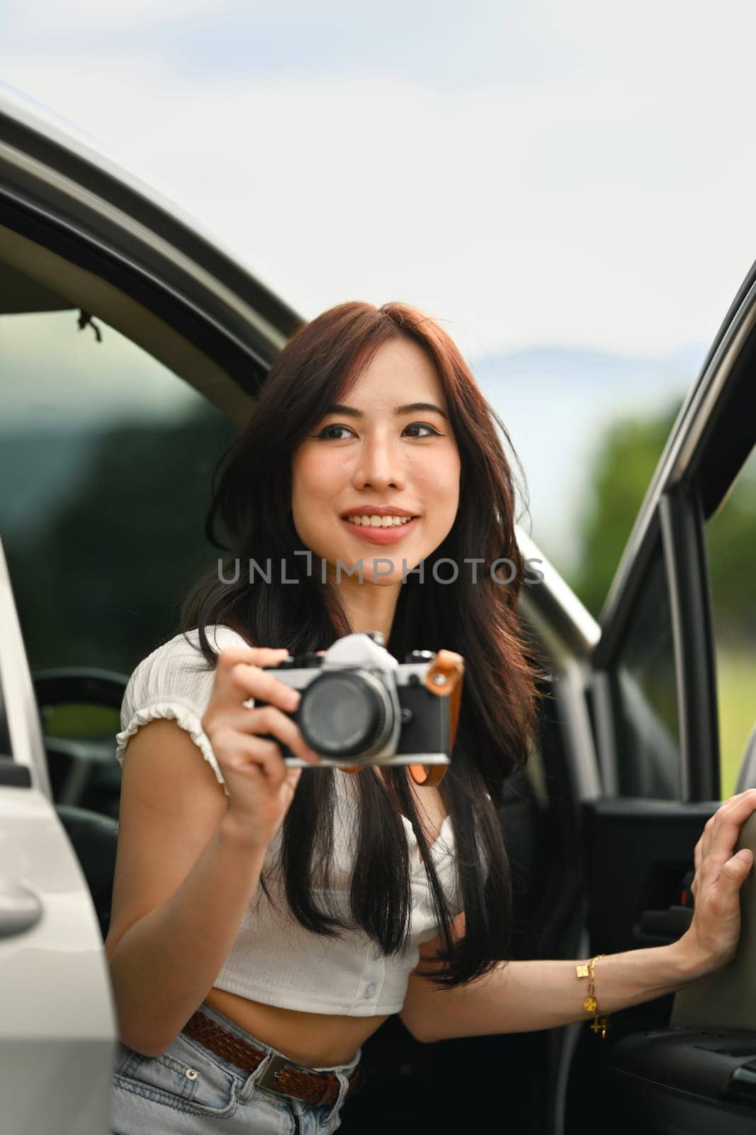 Charming millennial woman sitting in car with open door taking photos with vintage retro cameras during weekend road trip.