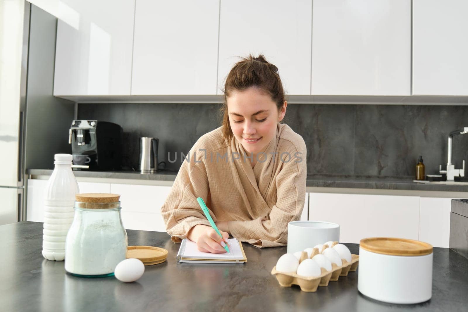 Attractive young cheerful girl baking at the kitchen, making dough, holding recipe book, having ideas.