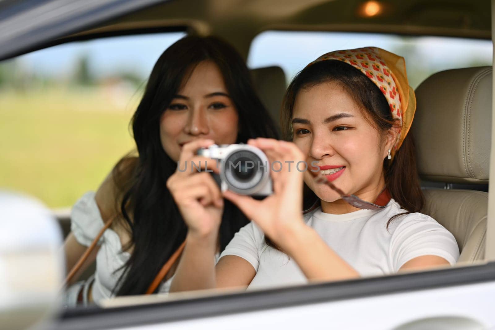 Two happy female friends taking photos from a car window with vintage retro cameras, enjoying weekend road trip together.