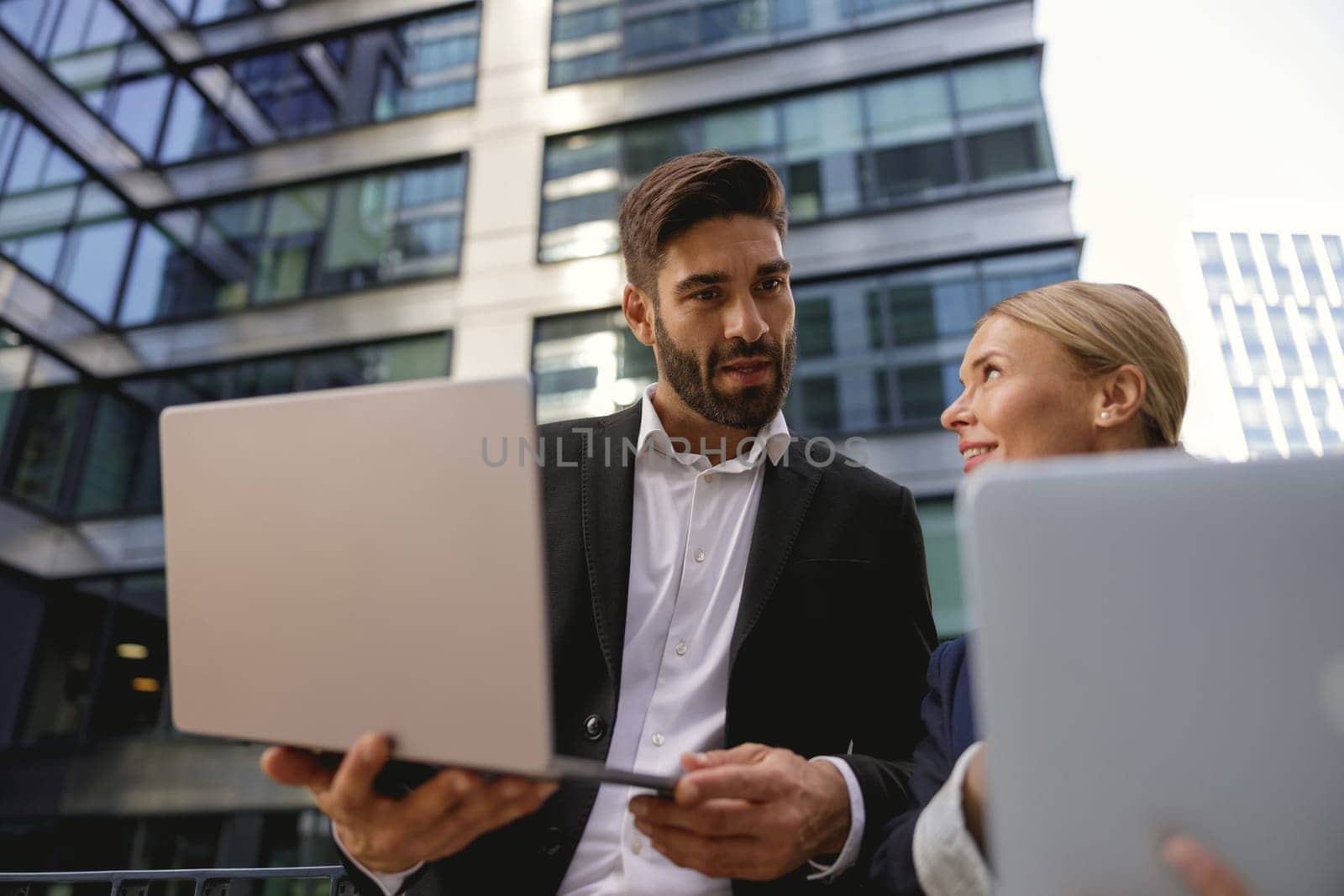 Two business people discuss biz issue while use laptop standing on modern office terrace