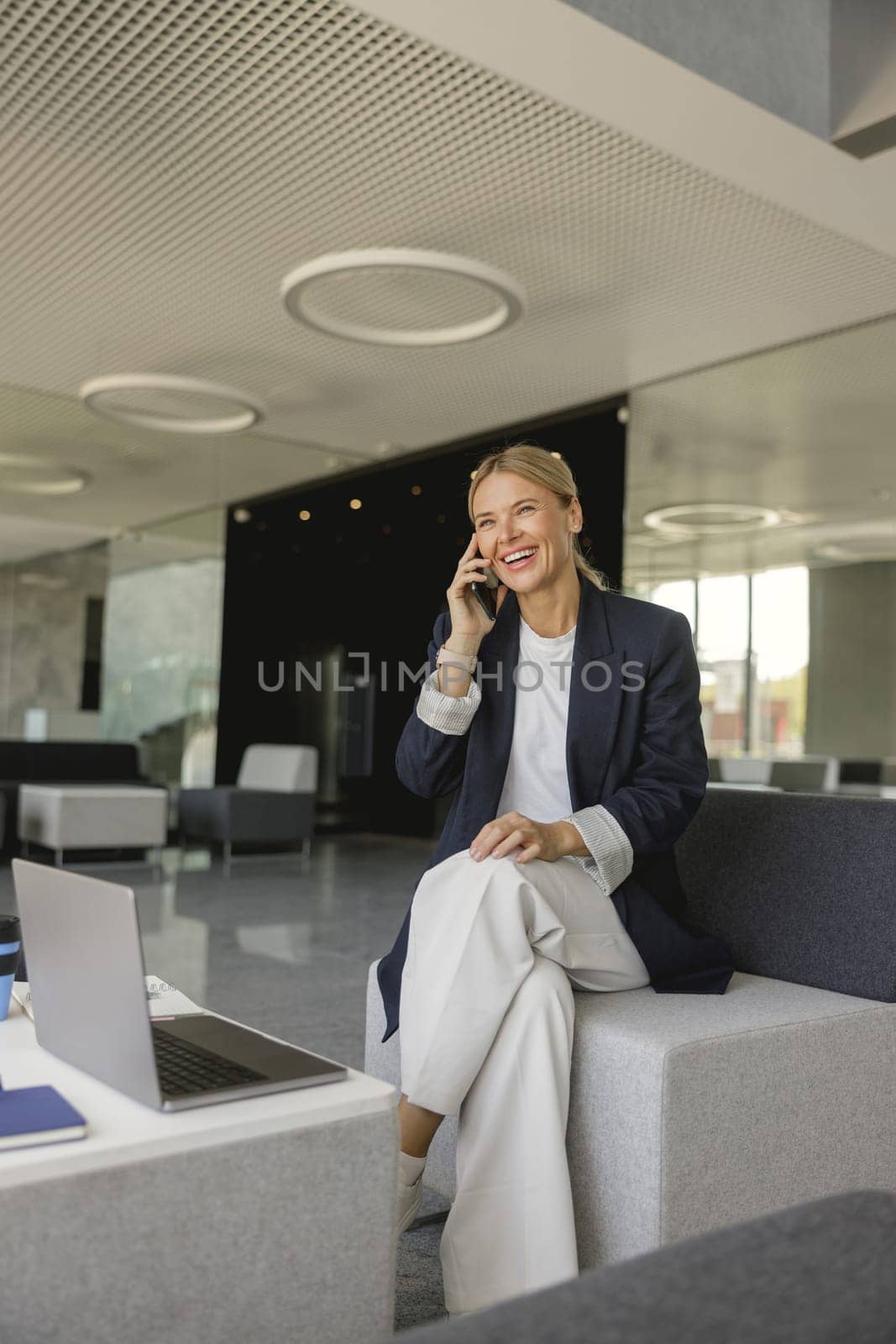 Smiling female entrepreneur talking phone sitting on background of modern office and looks away
