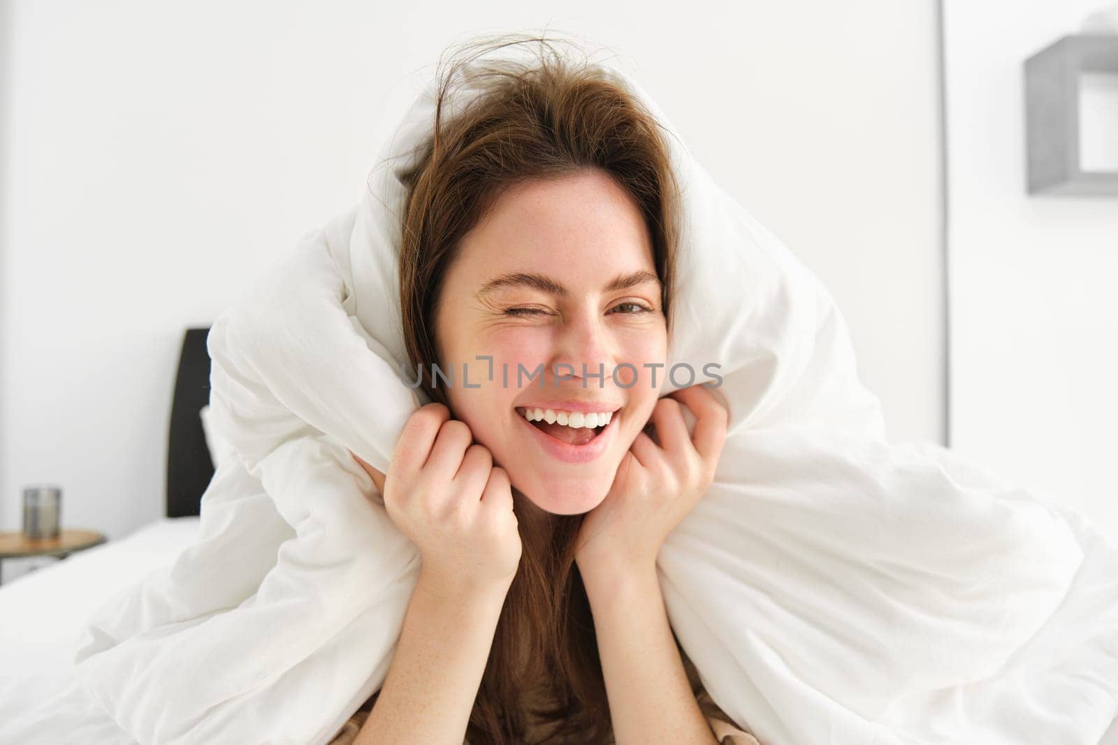 Portrait of gorgeous smiling woman, lying in bed covered in duvet, has messy hair, looking happy, relaxing in her bedroom, spending time in hotel room in morning.