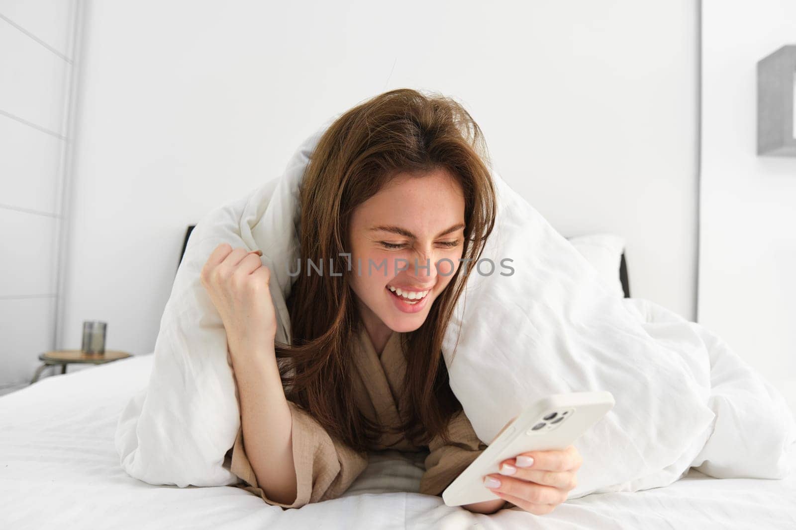 Woman with smartphone, feeling excitement, reading mobile phone and cheering, lying in bed under white blanket, using smartphone and looking happy in morning.