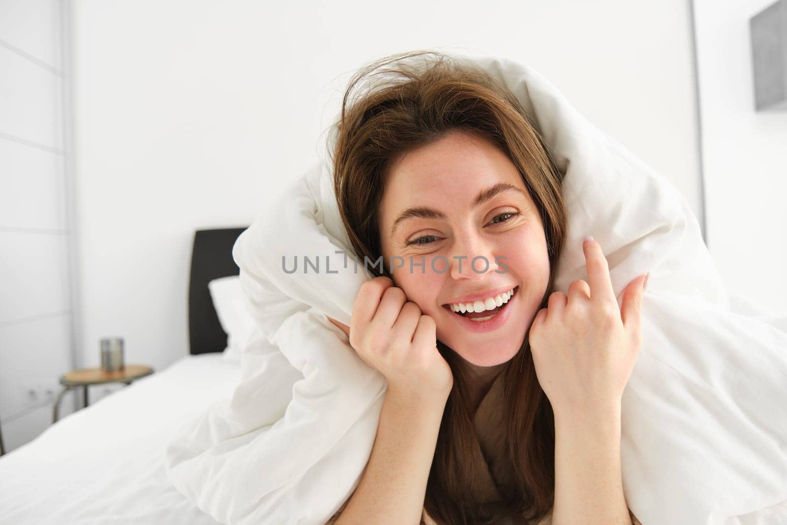 Portrait of beautiful brunette woman lying in bed, resting in her hotel room, covered in white blankets, laughing and smiling at camera.