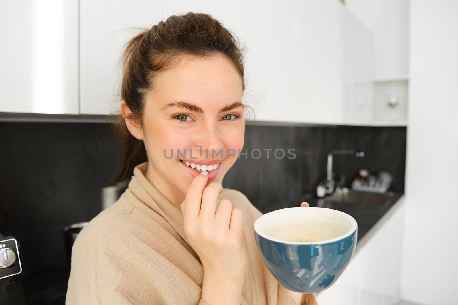 Close up portrait of attractive young woman in bathrobe, drinking coffee in the morning, holding cup of cappuccino and smiling, posing in the kitchen by Benzoix