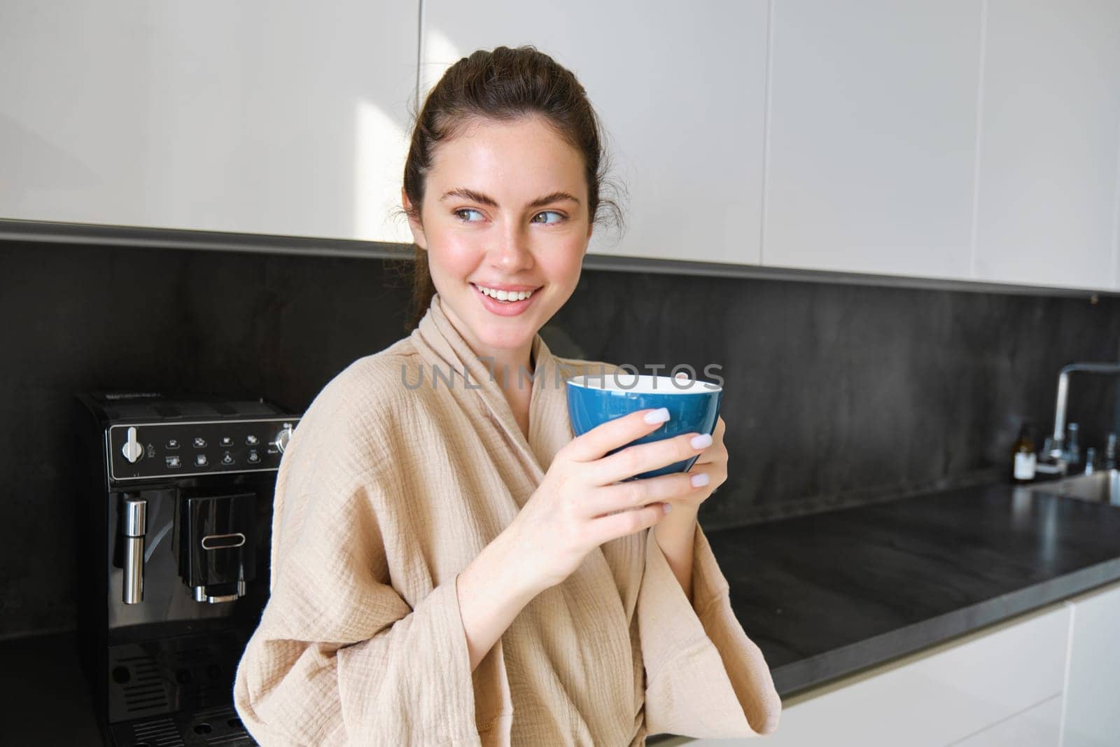 Lifestyle concept. Portrait of happy brunette woman in bathrobe, drinking coffee in the kitchen, having morning cuppa and smiling by Benzoix