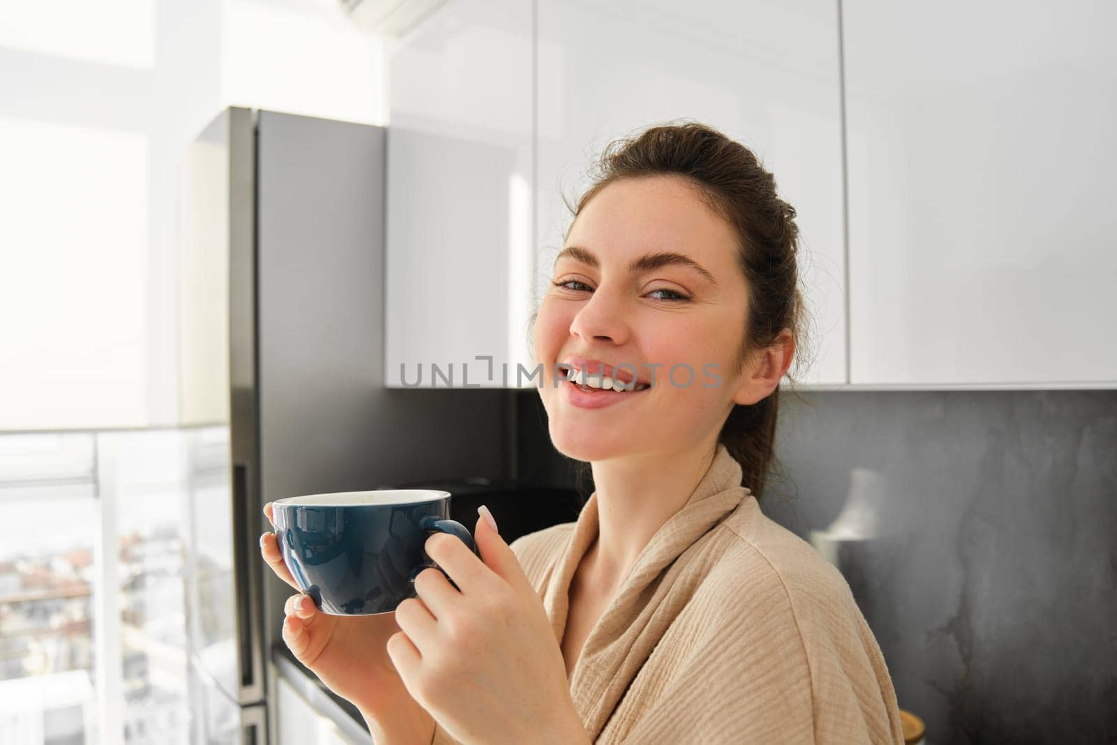 Portrait of happy young woman starts her morning with mug of coffee, drinking tea from cup, standing in the kitchen, smiling cheerfully.