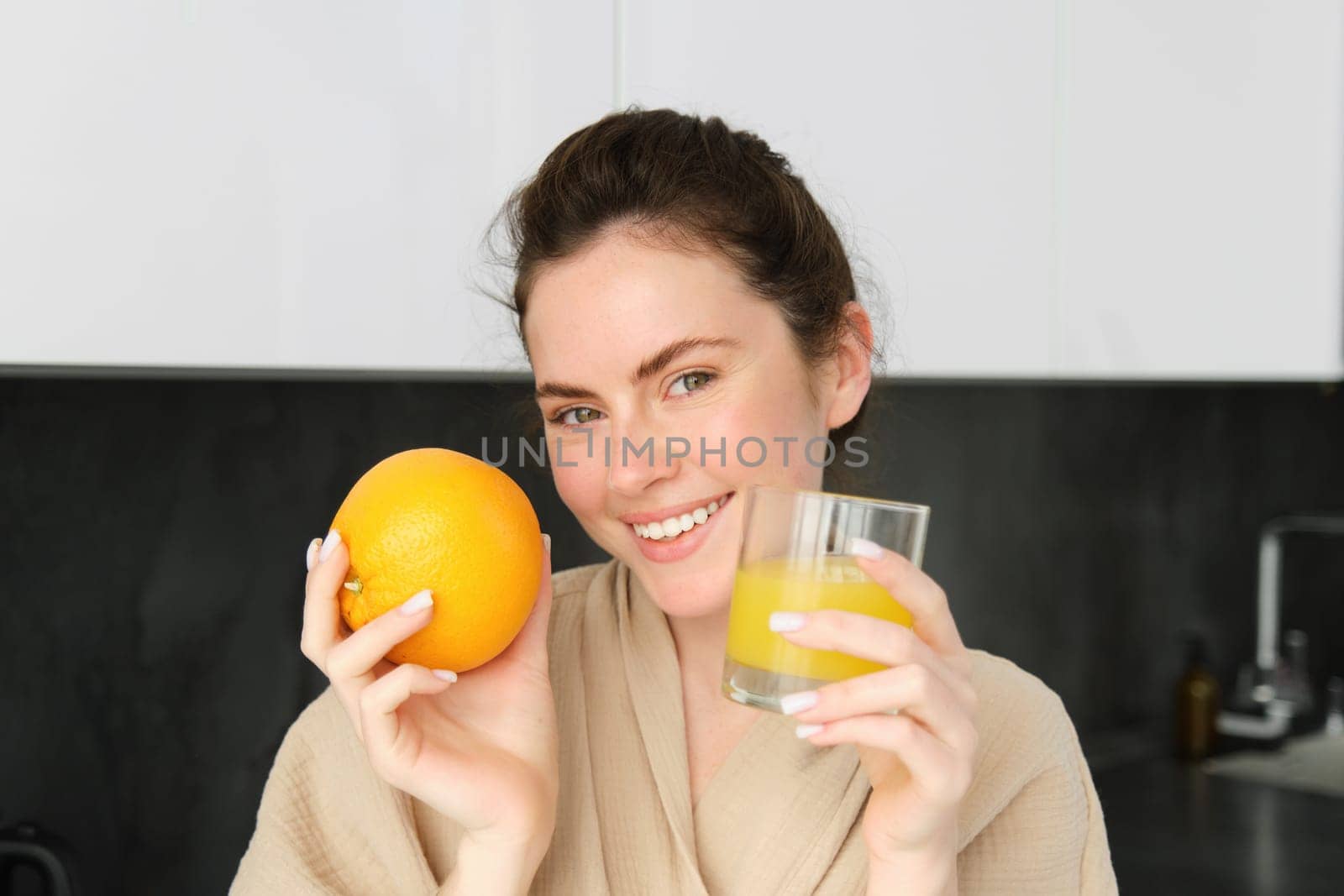 Image of good-looking healthy woman in bathrobe, drinking fresh juice, showing orange fruit, posing in kitchen by Benzoix