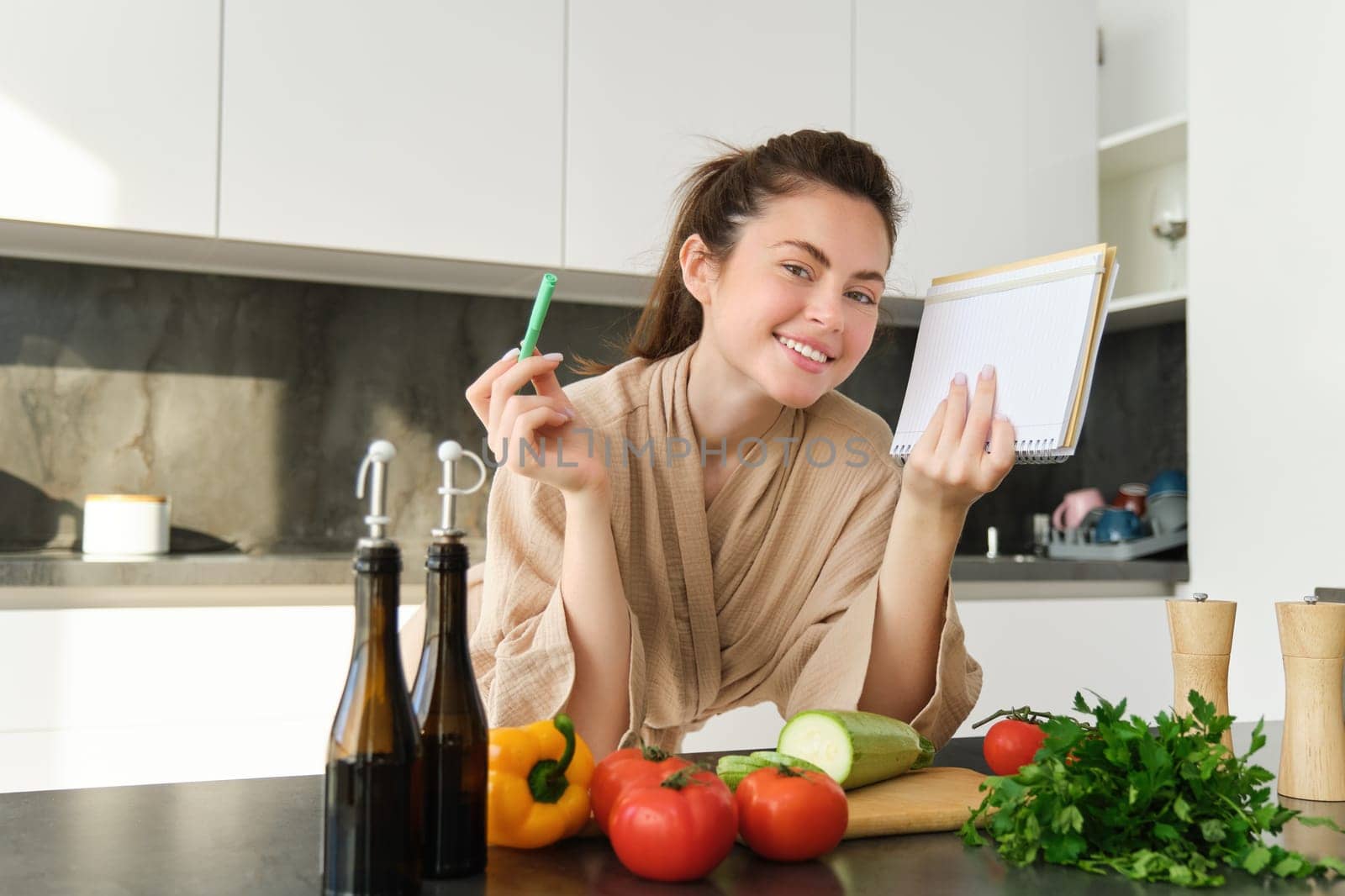 Portrait of good-looking, smiling girl with grocery list, holding notebook and reading recipe, cooking breakfast, making meal with vegetables, preparing salad.