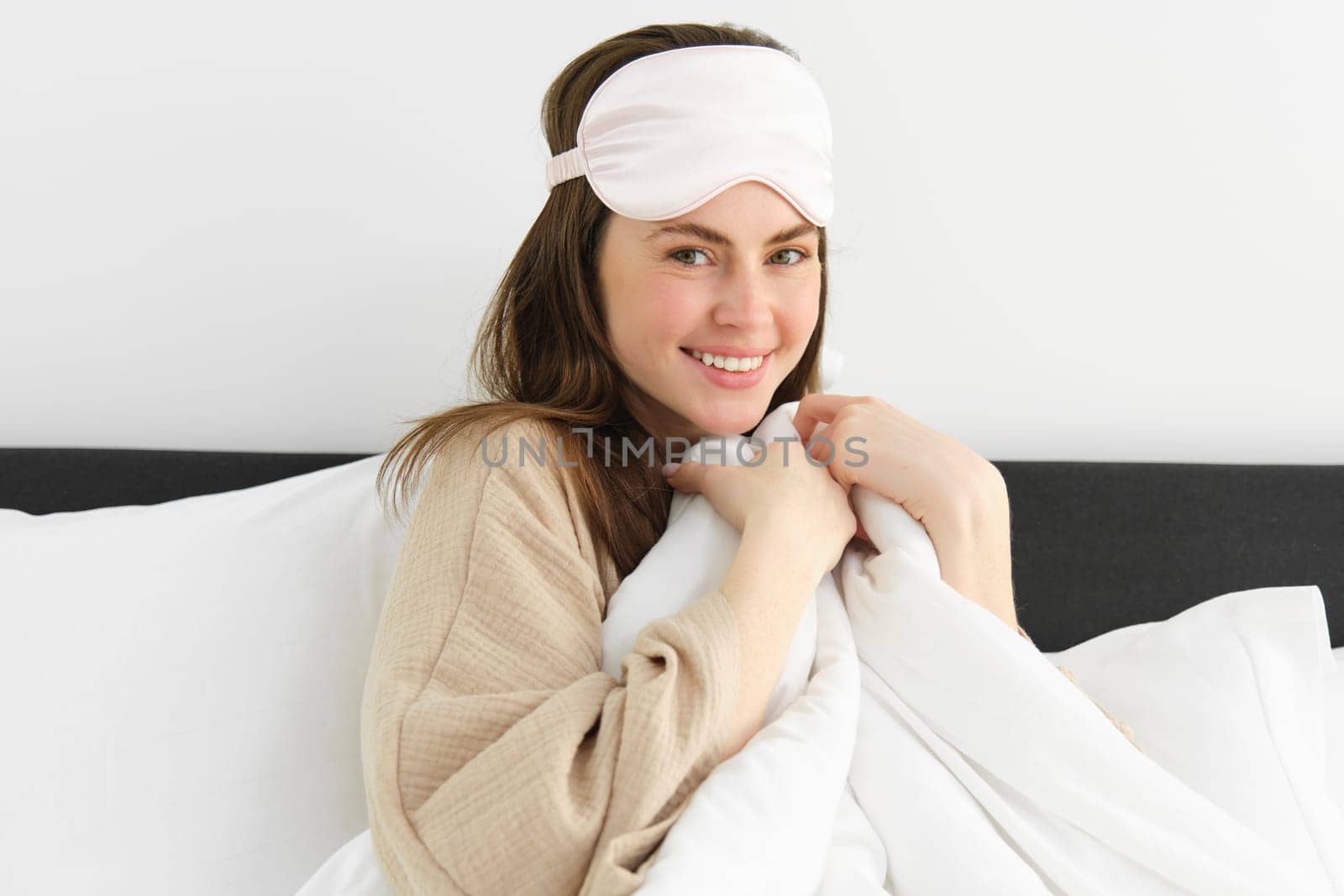 Pretty girl wakes up in morning, lying on pillow covered with white duvet, smiling at camera, wakes-up and looks happy.