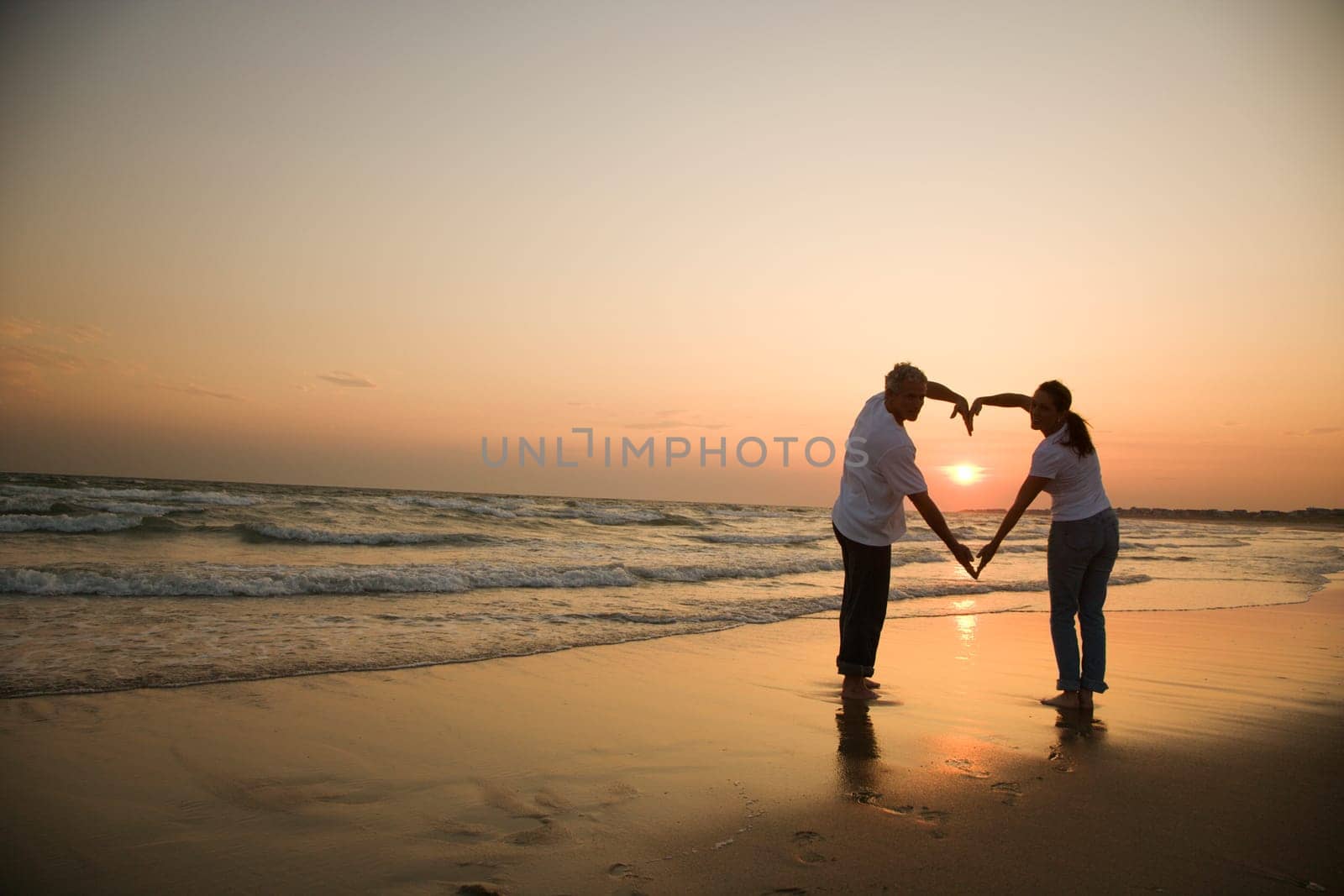 Mid-adult couple making heart shape with arms on beach at sunset.