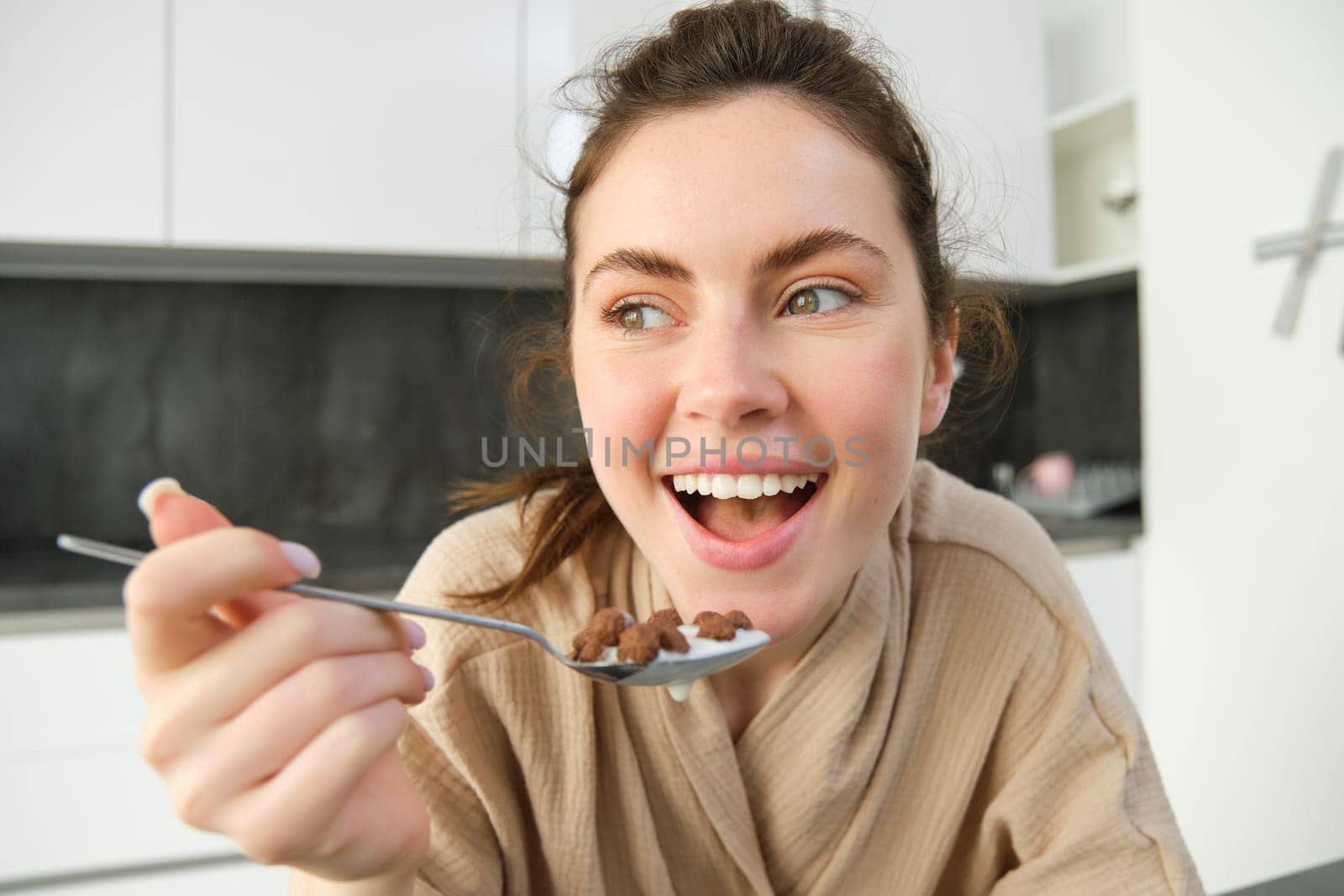 Close up portrait of happy smiling brunette in bathrobe, holding cereals in spoon, eating breakfast with milk and looking joyful, starting her day in morning.