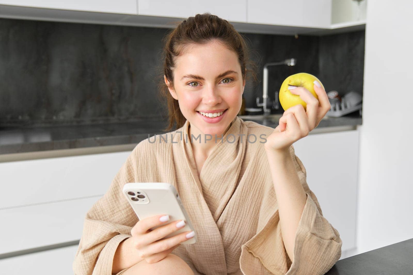 Close up portrait of happy young woman in bathrobe, sitting in the kitchen and using mobile phone, holding an apple, order fruits and vegetables online, using smartphone app for groceries delivery by Benzoix