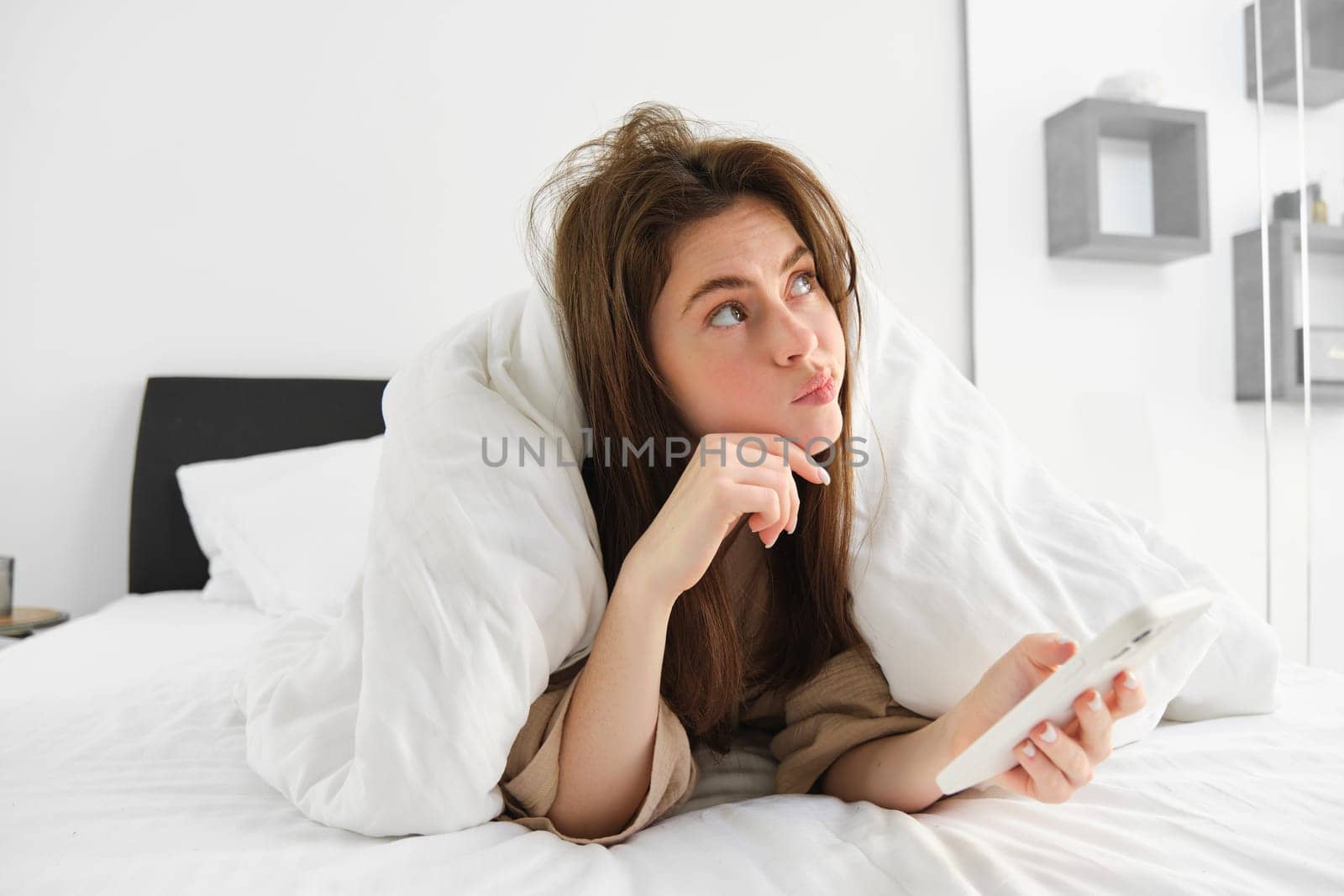 Photo of beautiful joyful woman in pyjama using mobile phone and smiling while lying on bed after sleep or nap.