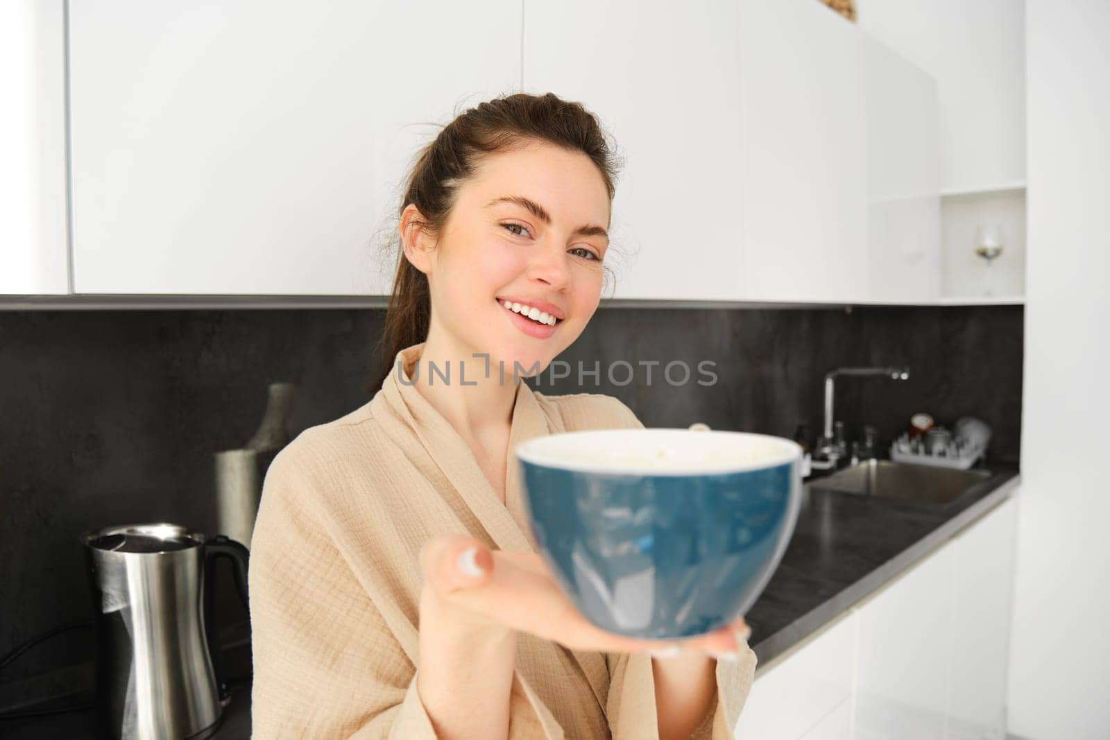 Portrait of good-looking young woman gives you cup of coffee, offering mug to you, standing in the kitchen, wearing bathrobe by Benzoix