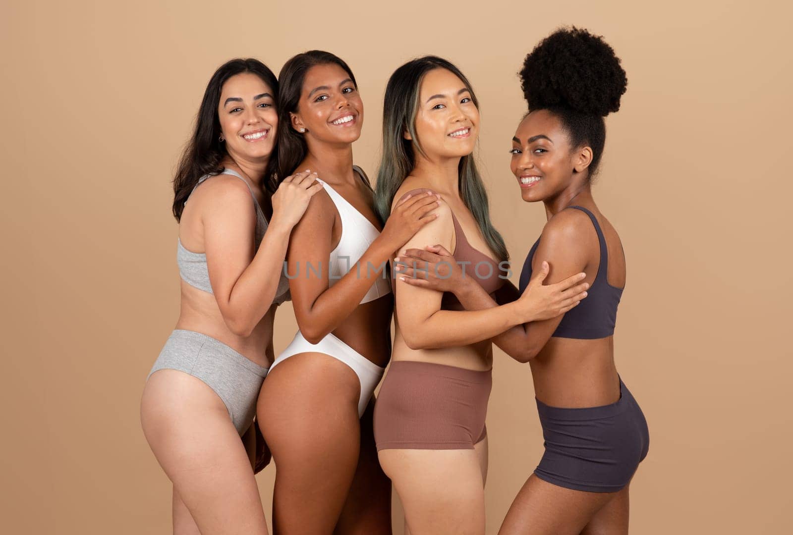 Four diverse young ladies confidently posing in their underwear, celebrating natural beauty and unity on beige studio background, looking and smiling at camera