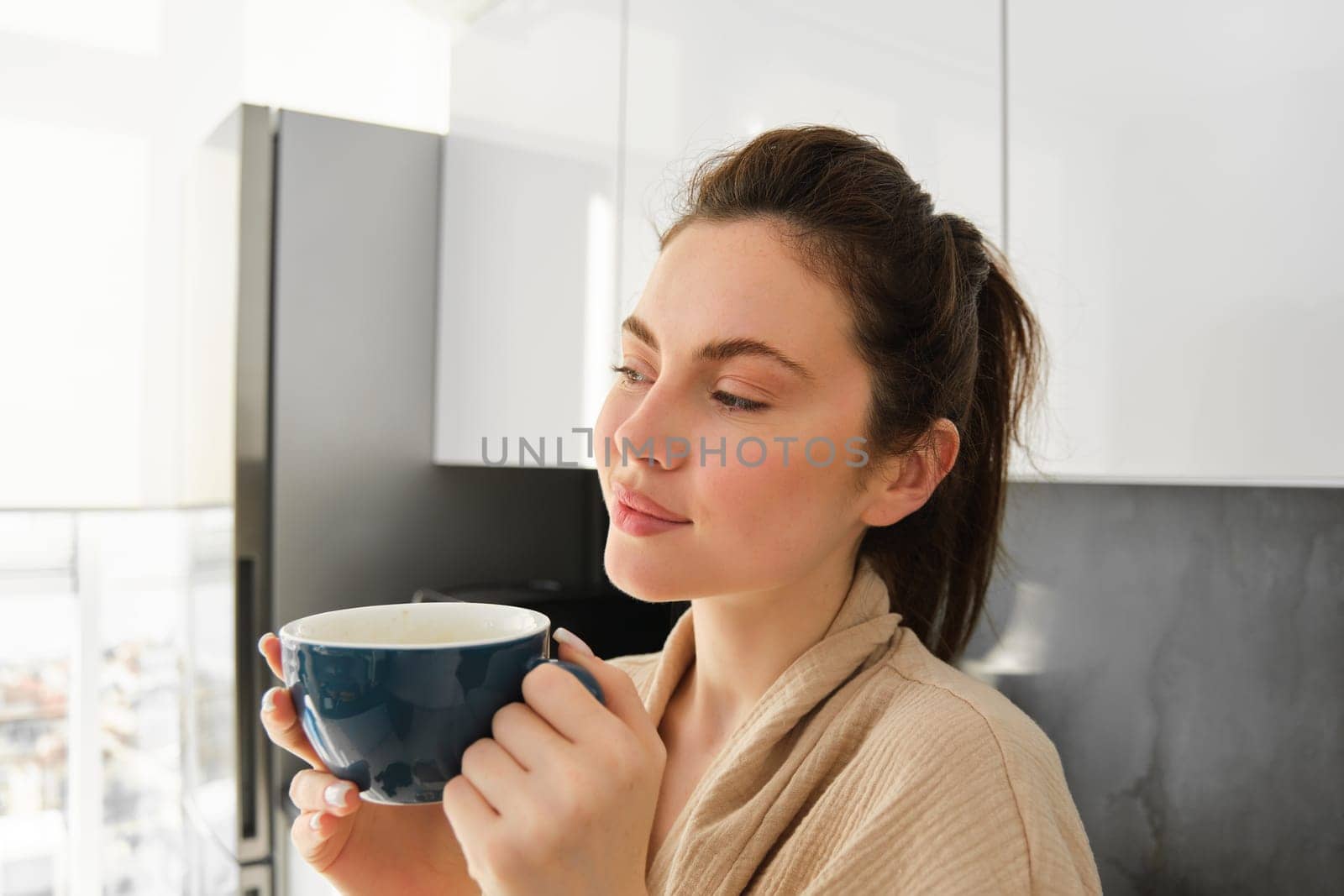 Daily routine and lifestyle. Young beautiful woman in bathrobe, standing in kitchen with cup of coffee, drinking tea, smiling and looking happy by Benzoix