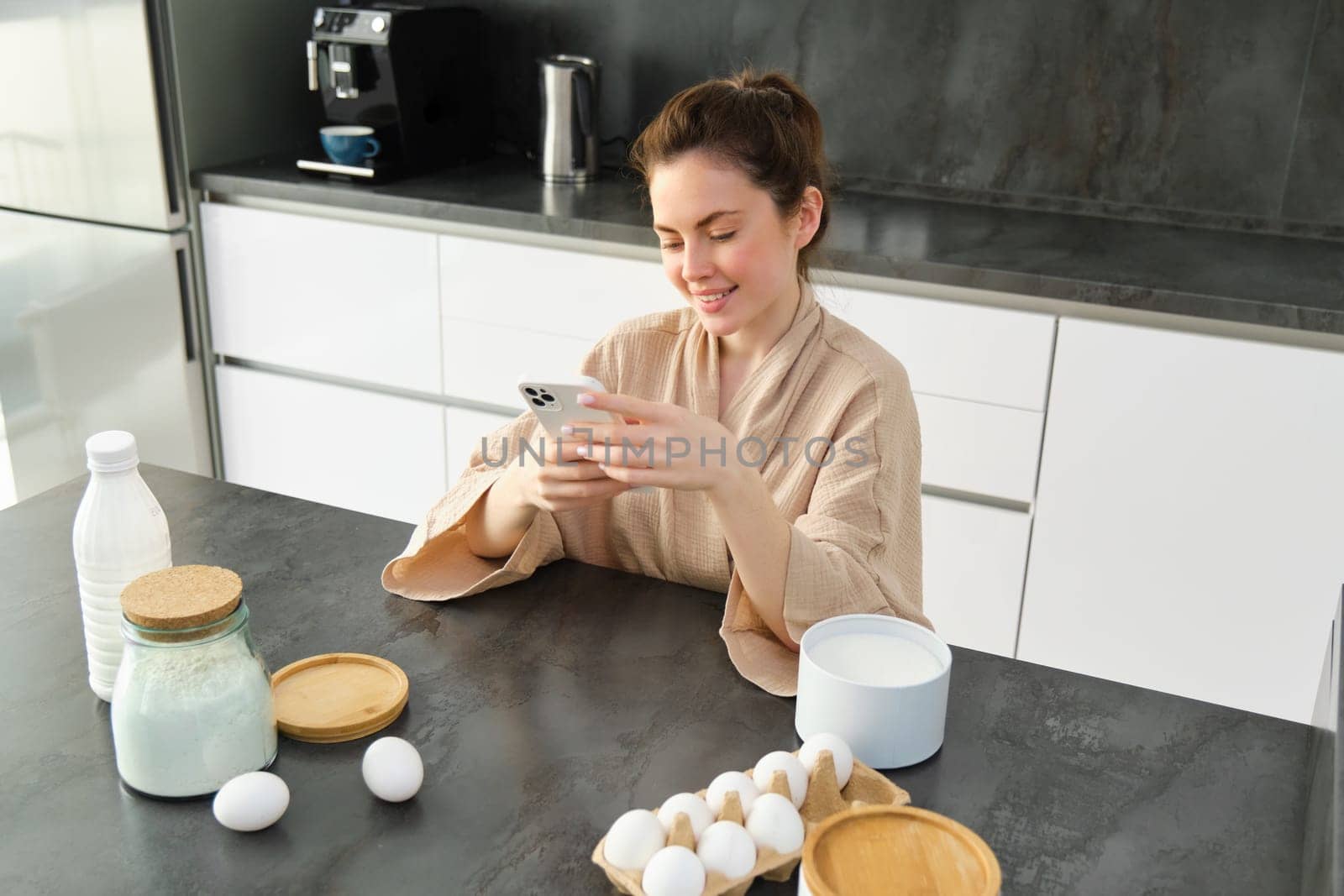 Attractive young cheerful girl baking at the kitchen, making dough, holding recipe book, having ideas.