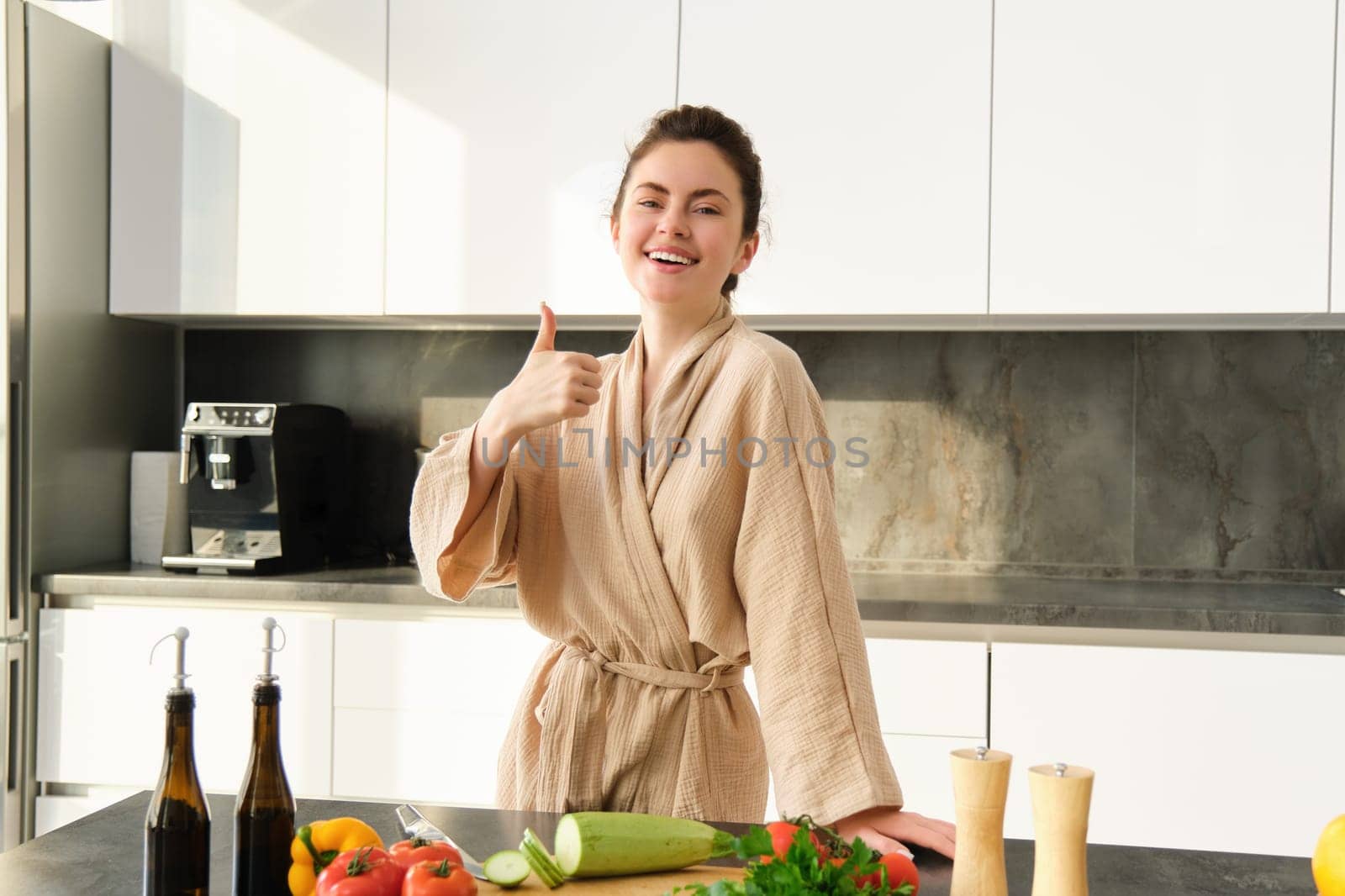 Portrait of happy young woman preparing vegetarian dinner, showing thumbs up, wife cooking in kitchen and smiling, recommending product, chopping vegetables for healthy salad by Benzoix