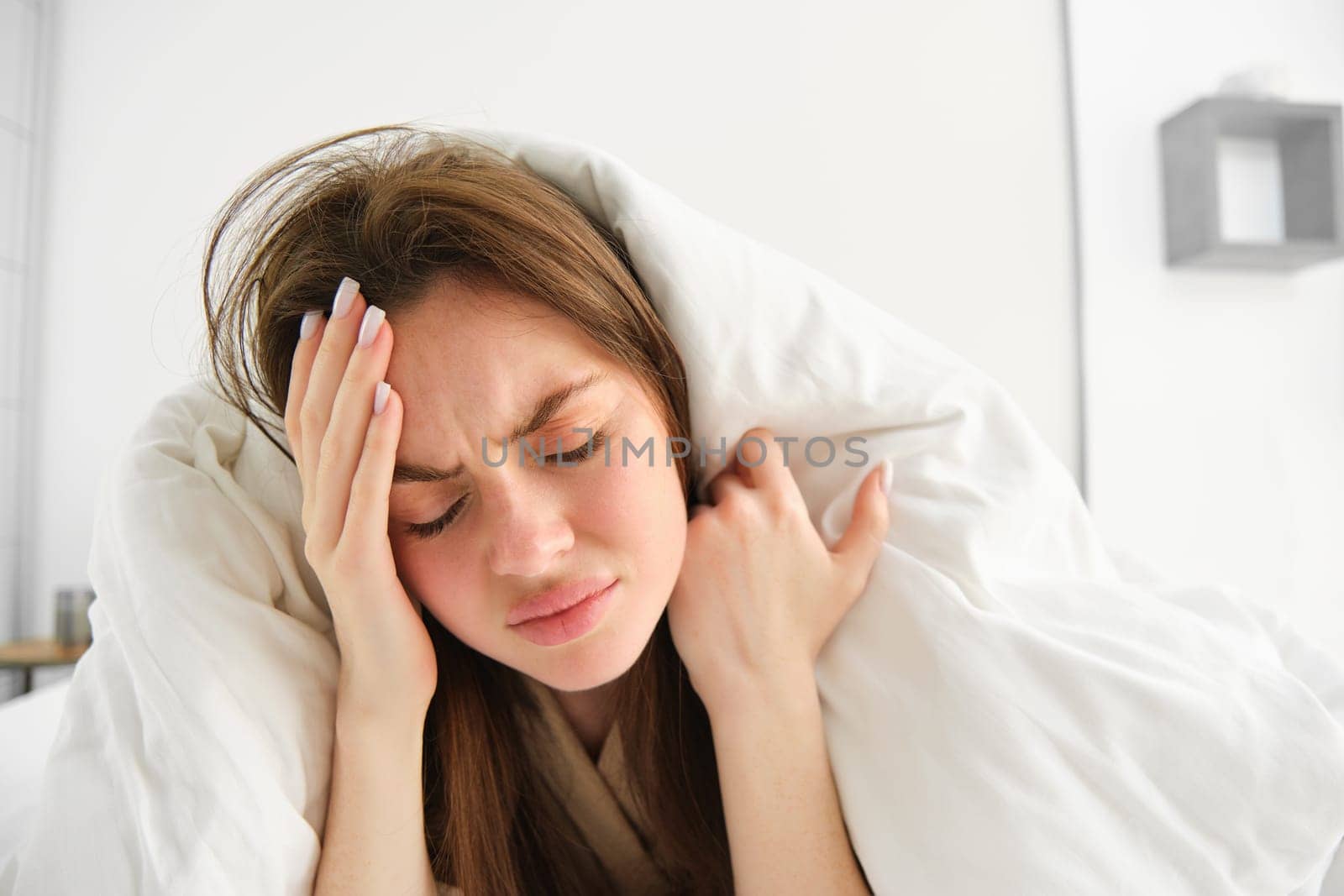 Close up of woman feeling unwell at home, lying in bed under white blankets and frowning, touching head, has headache, migraine, recovering from covid by Benzoix