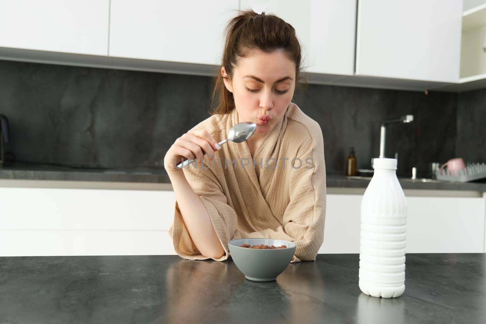 Portrait of young beautiful woman in bathrobe, eating cereals for breakfast, leans on kitchen worktop, looking at her morning meal.