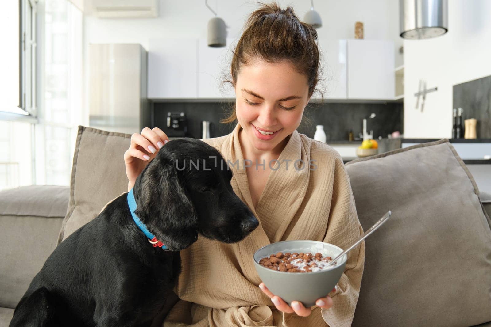 Happy young woman in bathrobe, enjoys spending time with her dog at home, eating cereals, having breakfast with puppy, sitting on sofa.