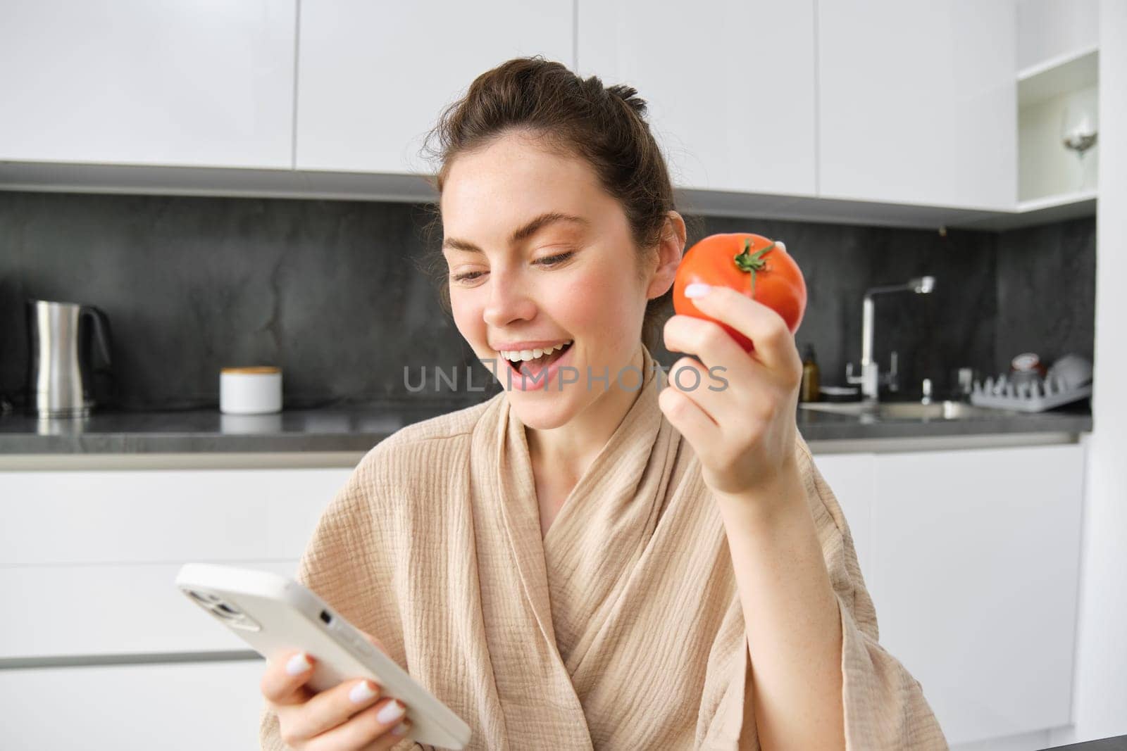 Close up portrait of beautiful young woman in the kitchen, holding tomato and smartphone, buys fresh vegetables online, orders groceries delivery on mobile phone application by Benzoix