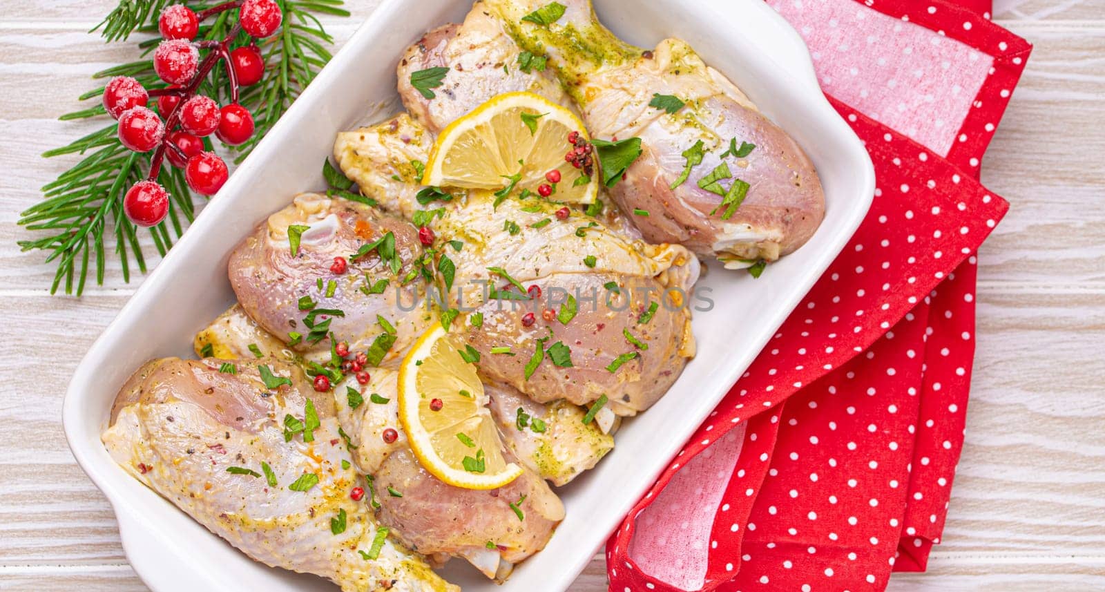 Preparing festive Christmas Eve meal with marinated chicken drumsticks. Raw chicken legs with herbs, lemon in white ceramic casserole top view on light wooden kitchen table with Christmas decoration.