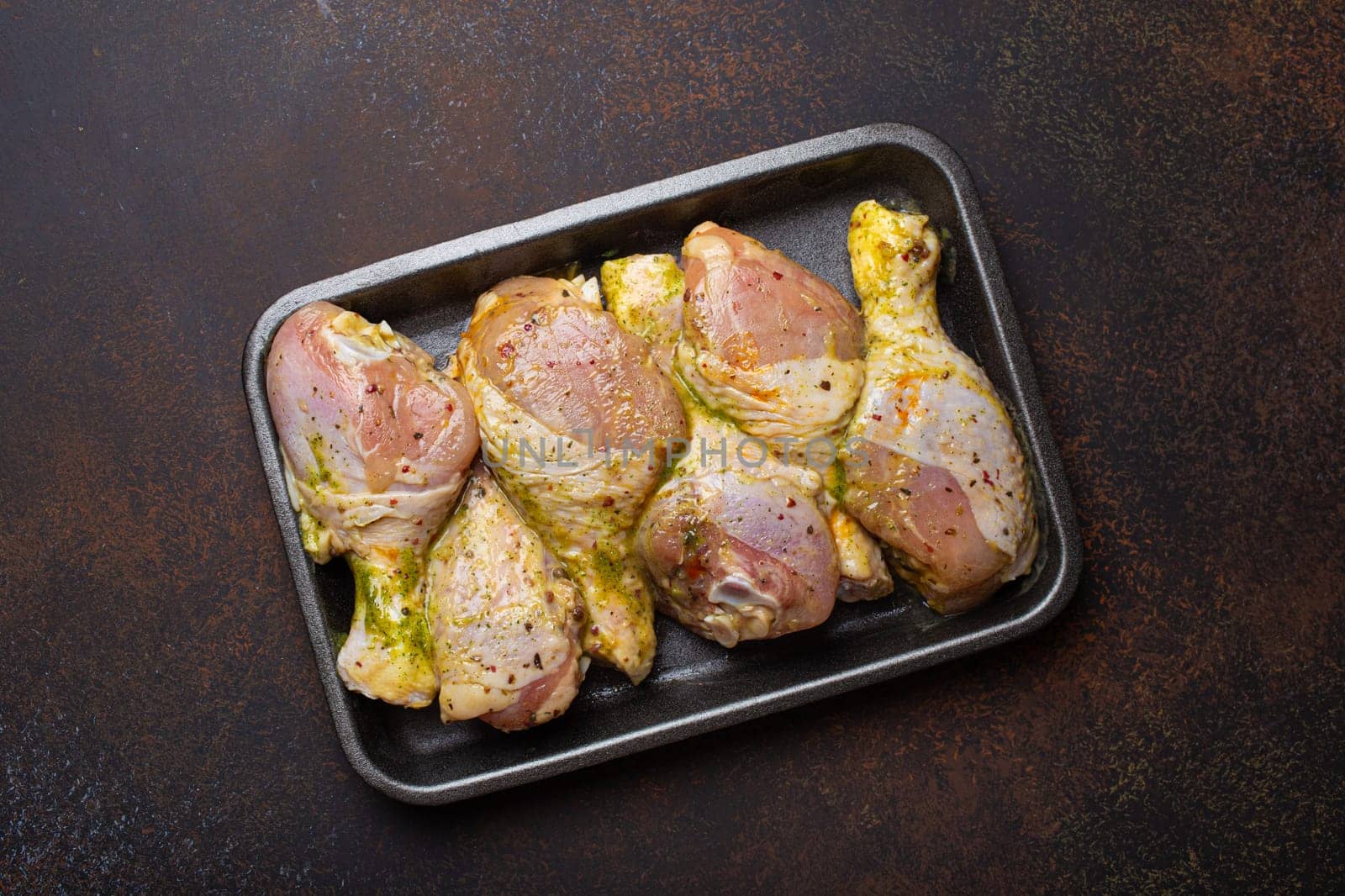 Raw uncooked chicken legs in green marinade with seasonings in black plastic container top view on dark rustic background. Preparing healthy meal with marinated chicken drumsticks.