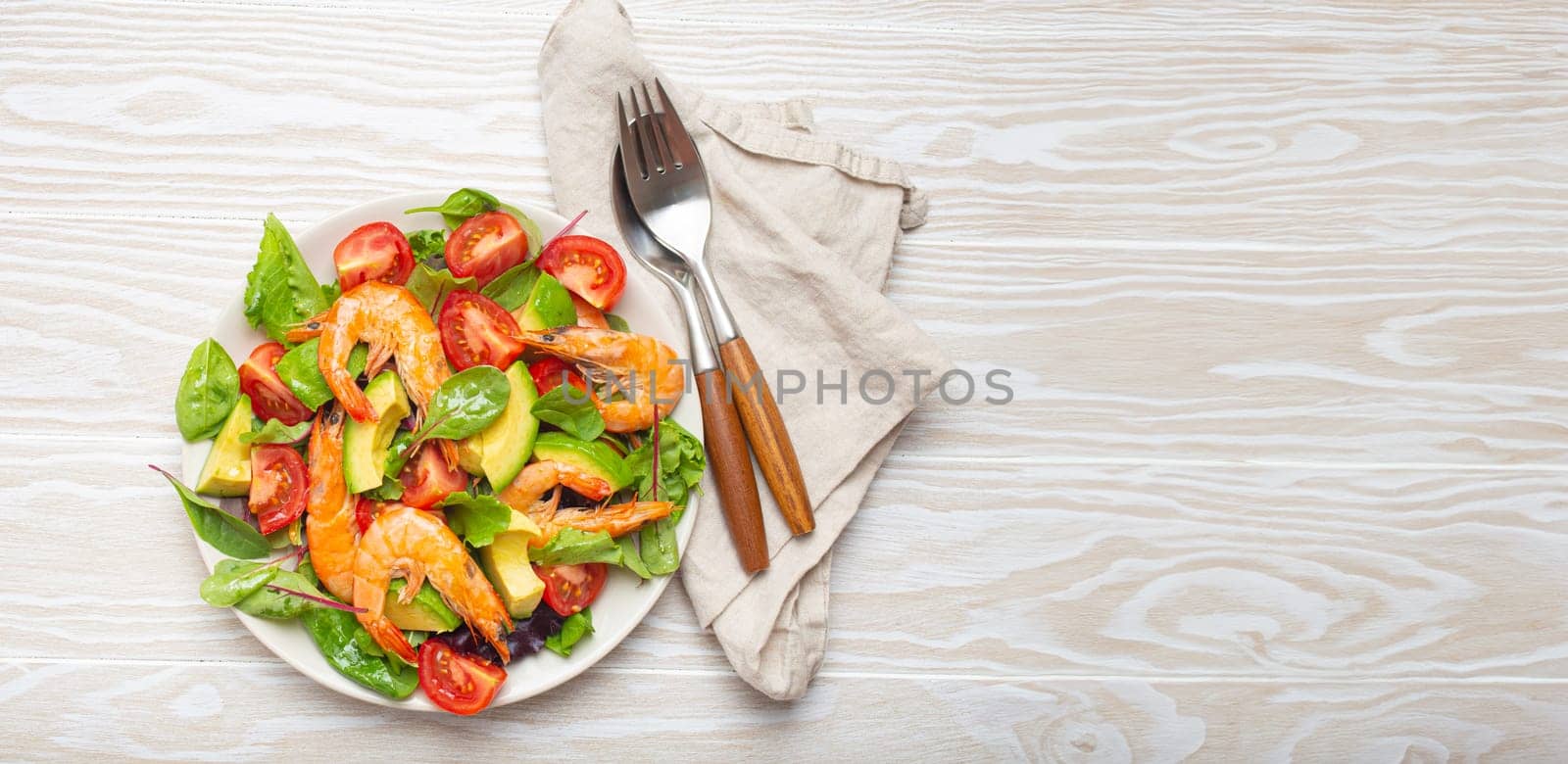 Healthy salad with grilled shrimps, avocado, cherry tomatoes and green leaves on white plate with cutlery on white wooden rustic background top view. Clean eating, nutrition and dieting, copy space..