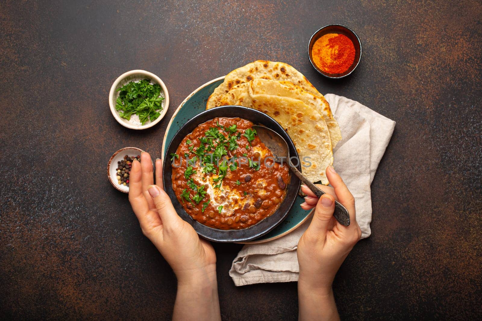 Female hands holding a bowl and eating traditional Indian Punjabi dish Dal makhani with lentils and beans served with naan flat bread, fresh cilantro on brown concrete rustic table top view by its_al_dente