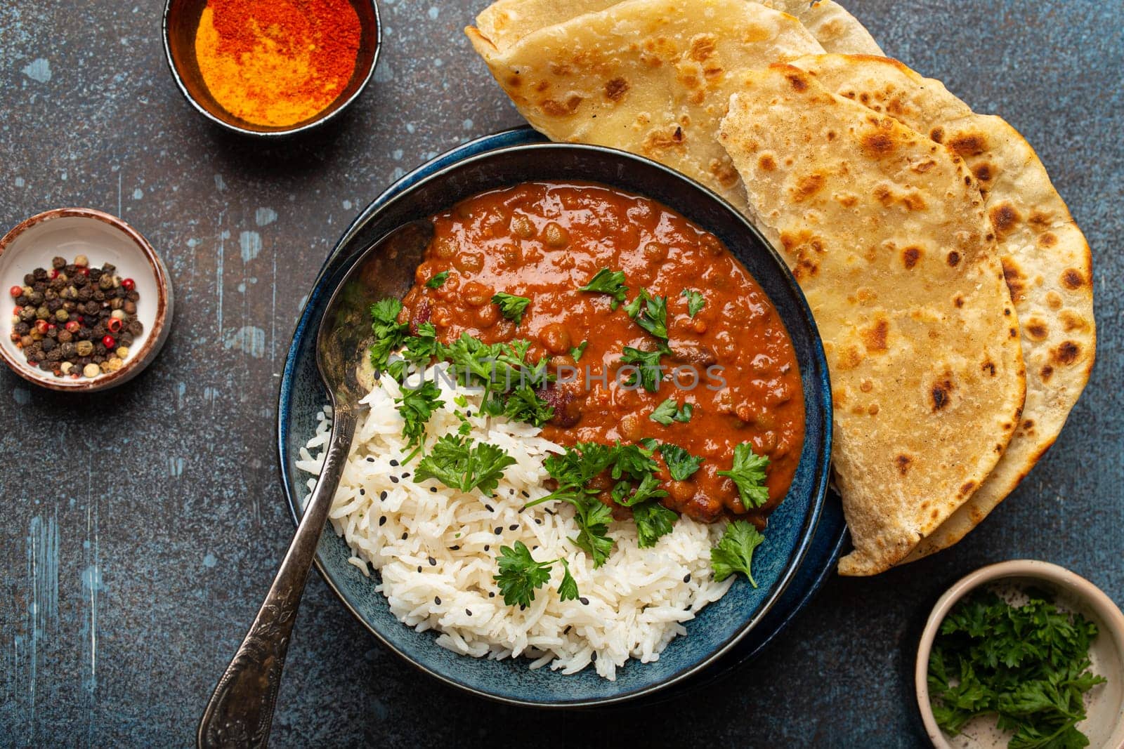 Traditional Indian Punjabi dish Dal makhani with lentils and beans in black bowl served with basmati rice, naan flat bread, fresh cilantro and spoon on blue concrete rustic table top view by its_al_dente