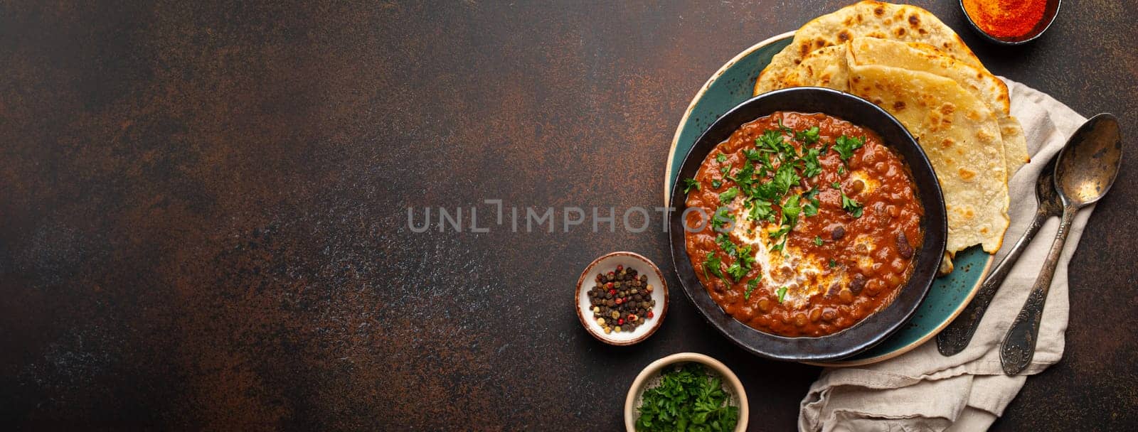 Traditional Indian Punjabi dish Dal makhani with lentils and beans in black bowl served with naan flat bread, fresh cilantro and two spoons on brown concrete rustic table top view. Space for text.