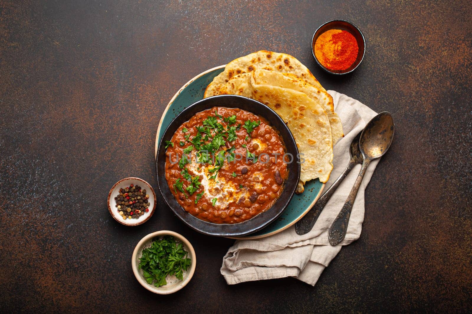 Traditional Indian Punjabi dish Dal makhani with lentils and beans in black bowl served with naan flat bread, fresh cilantro and two spoons on brown concrete rustic table top view. Space for text.