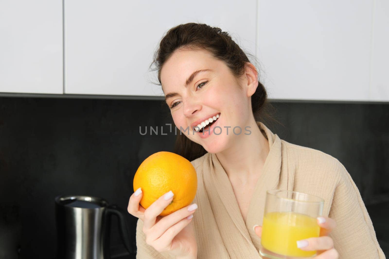 Portrait of attractive young modern woman, posing in kitchen, holding glass of juice and an orange, laughing and talking to someone aside.