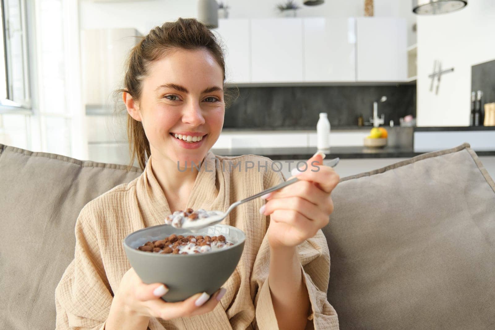 Relaxing morning at home. Young beautiful woman in bathrobe, eating cereals on sofa, having her breakfast in living room.