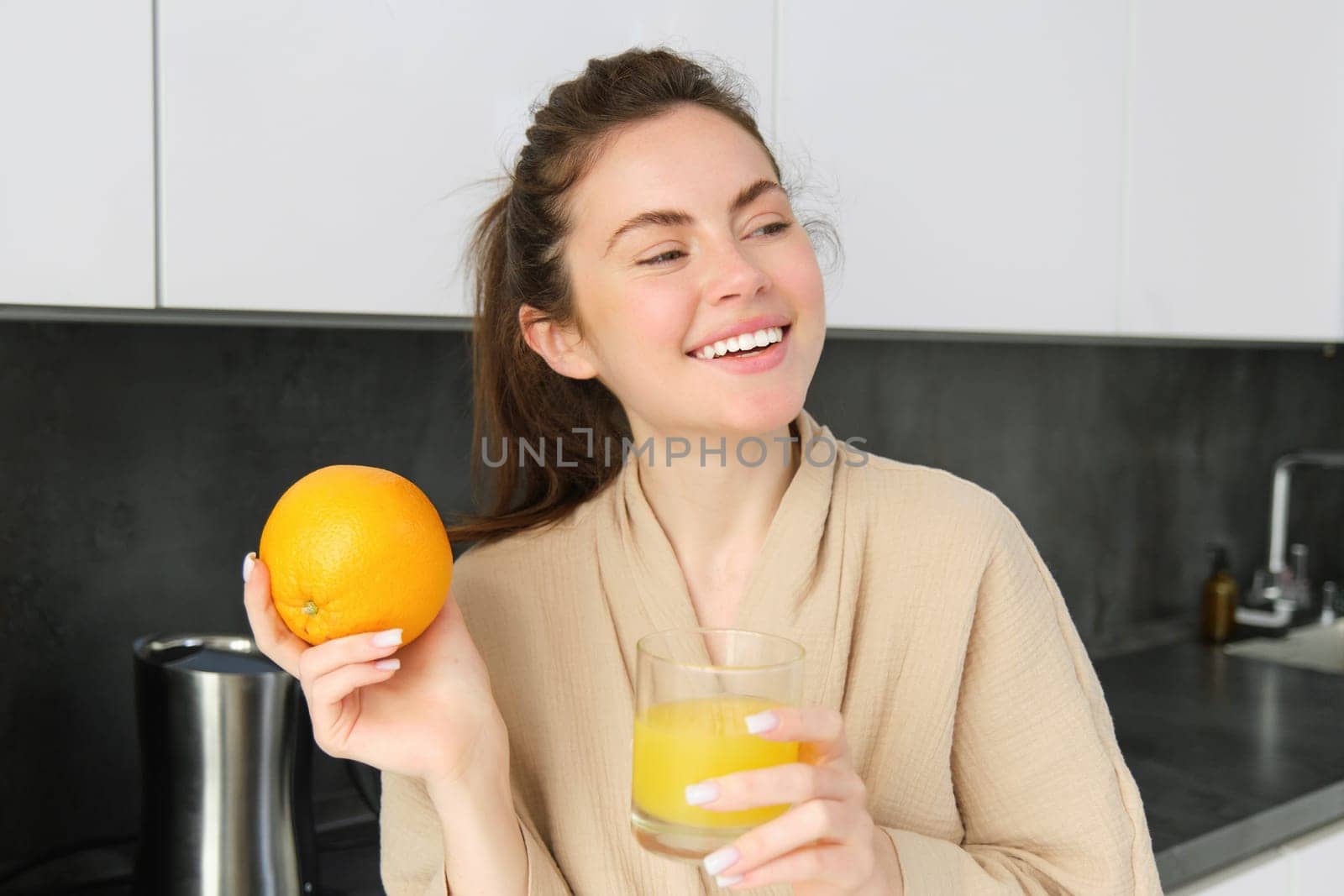 Image of good-looking healthy woman in bathrobe, drinking fresh juice, showing orange fruit, posing in kitchen.