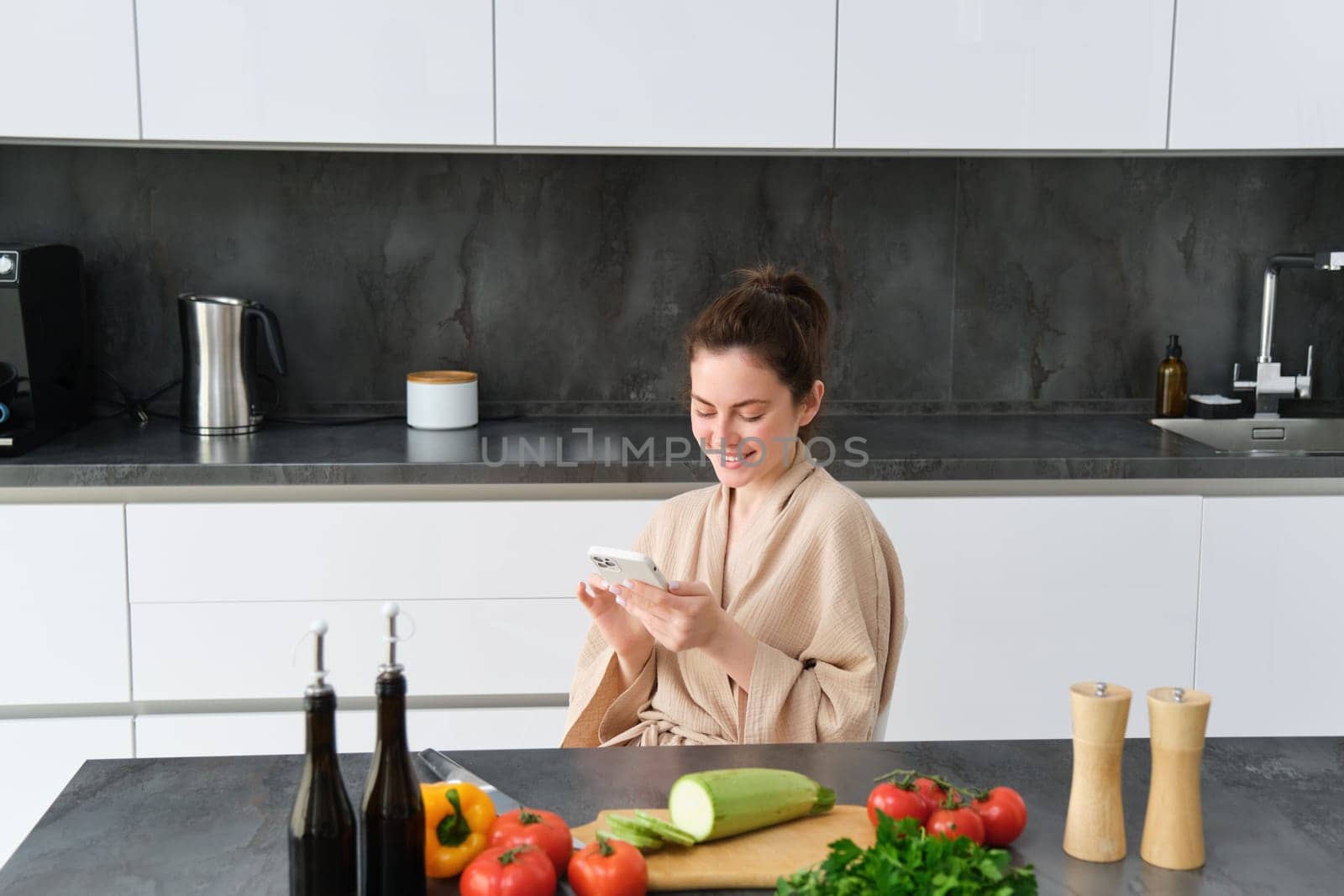 Woman watching recipe on smartphone, sitting in the kitchen with vegetables and chopping board, preparing dinner, healthy salad, cooking at home, wearing bathrobe by Benzoix
